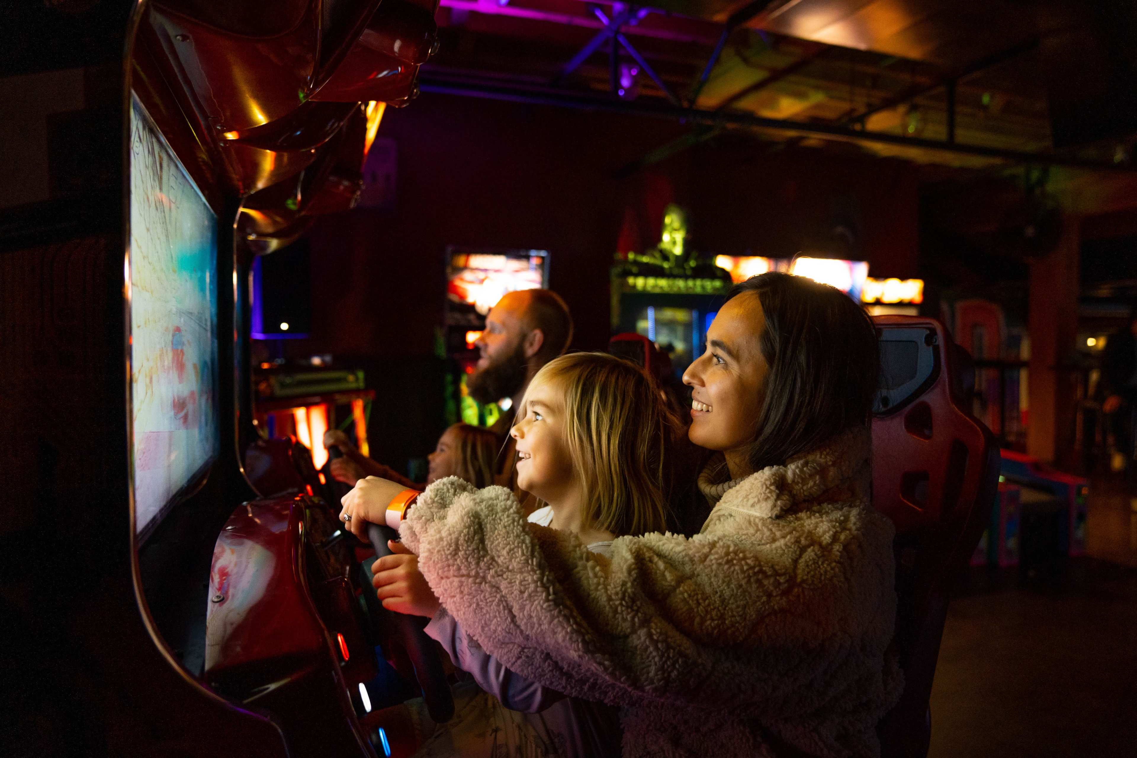 Two children and an adult are playing an arcade racing game together in a colorful game room.