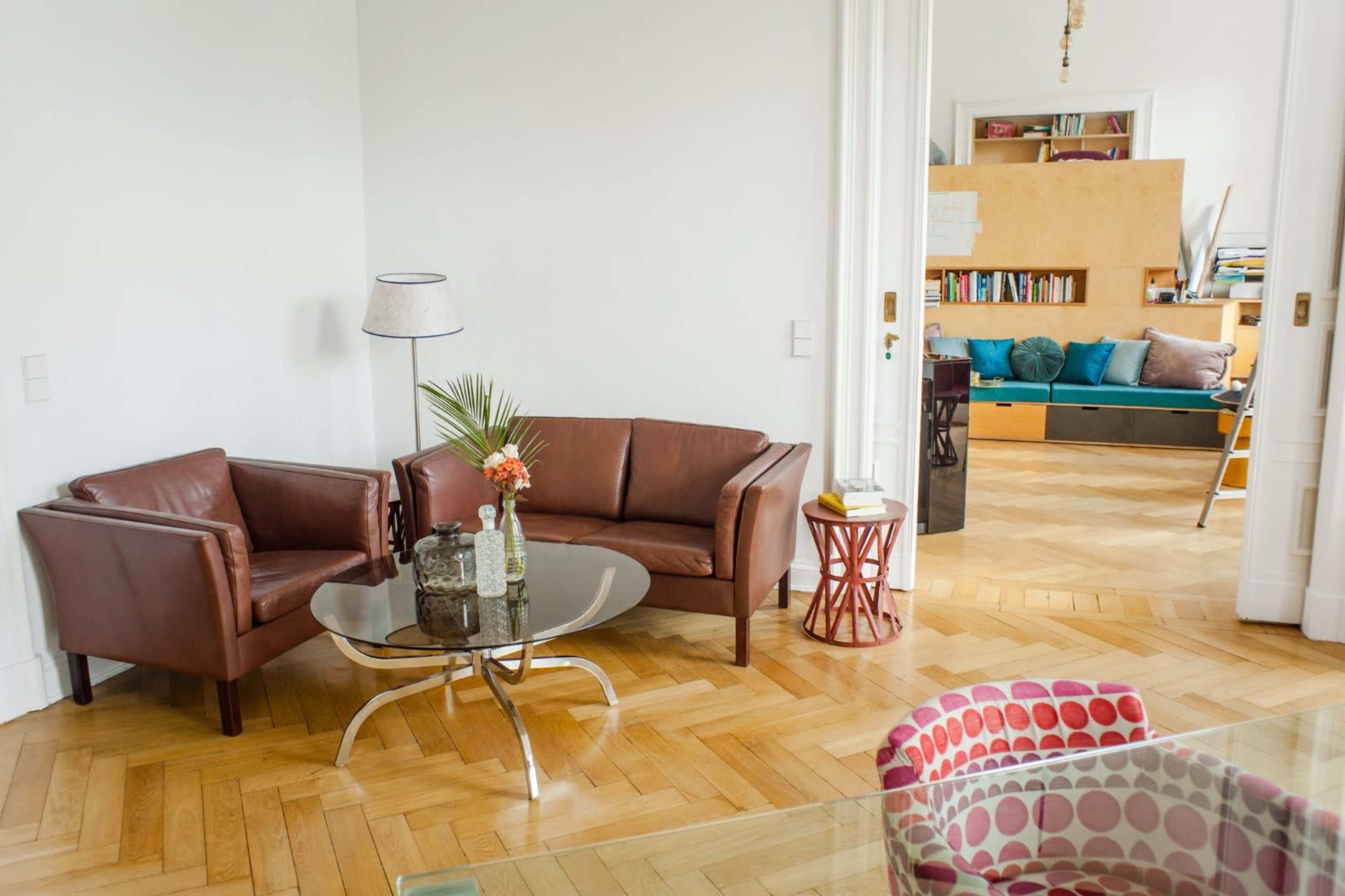 A living room with two brown leather chairs, a glass coffee table, and a colorful patterned chair, accented by wooden flooring and a visible bookshelf in the background.