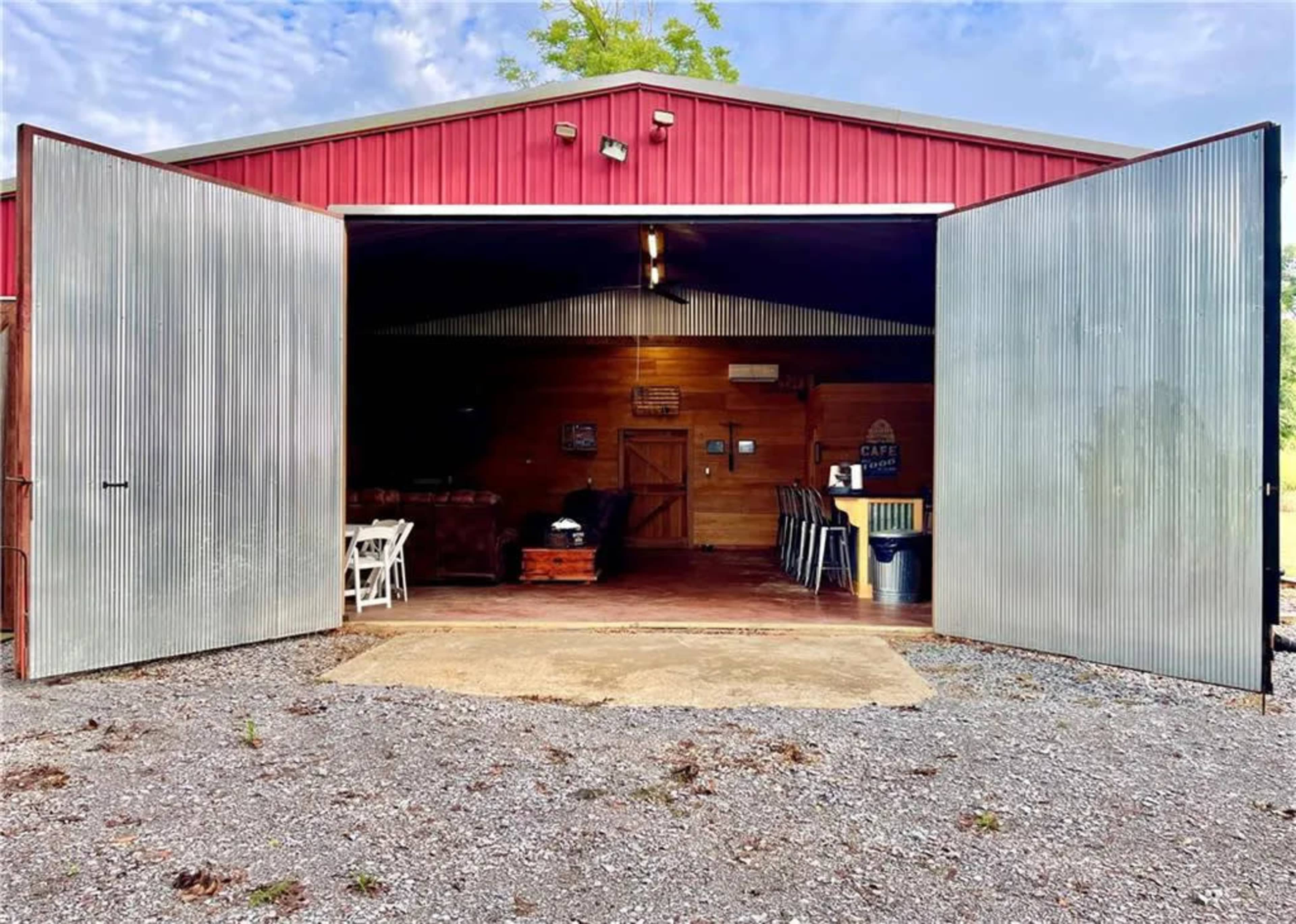 A red barn with large, open metal doors reveals a cluttered interior space with wooden furnishings and tables.