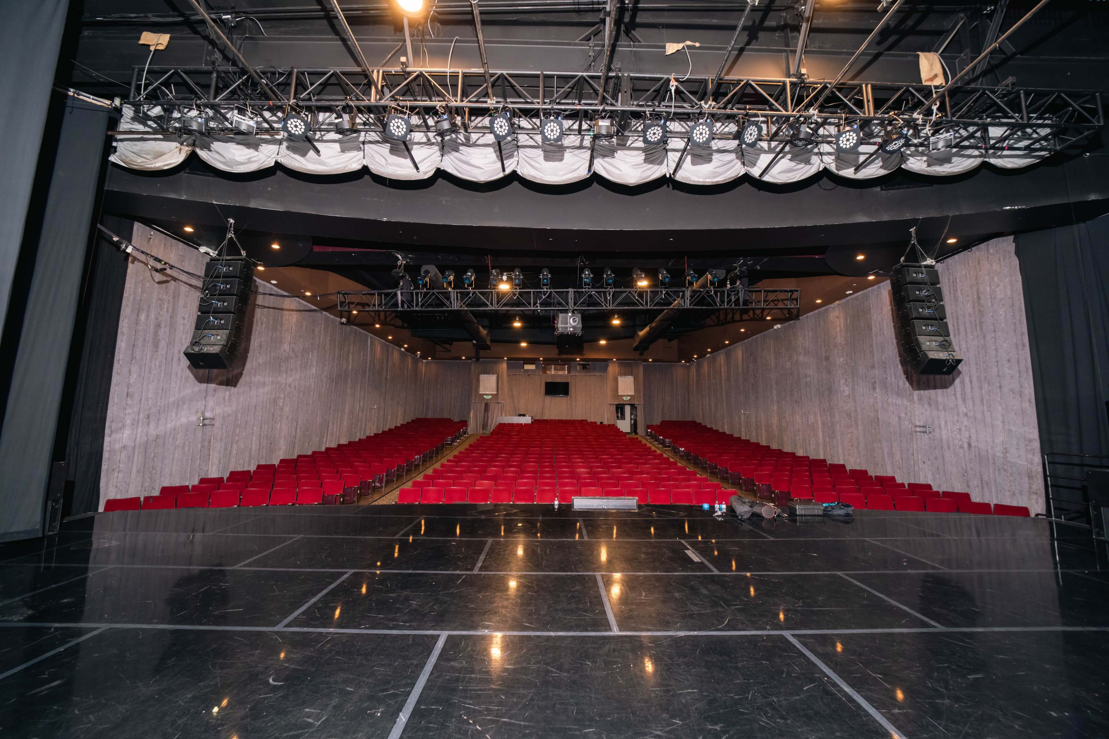 An empty theater stage with red seating rows and a grid of stage lights overhead.