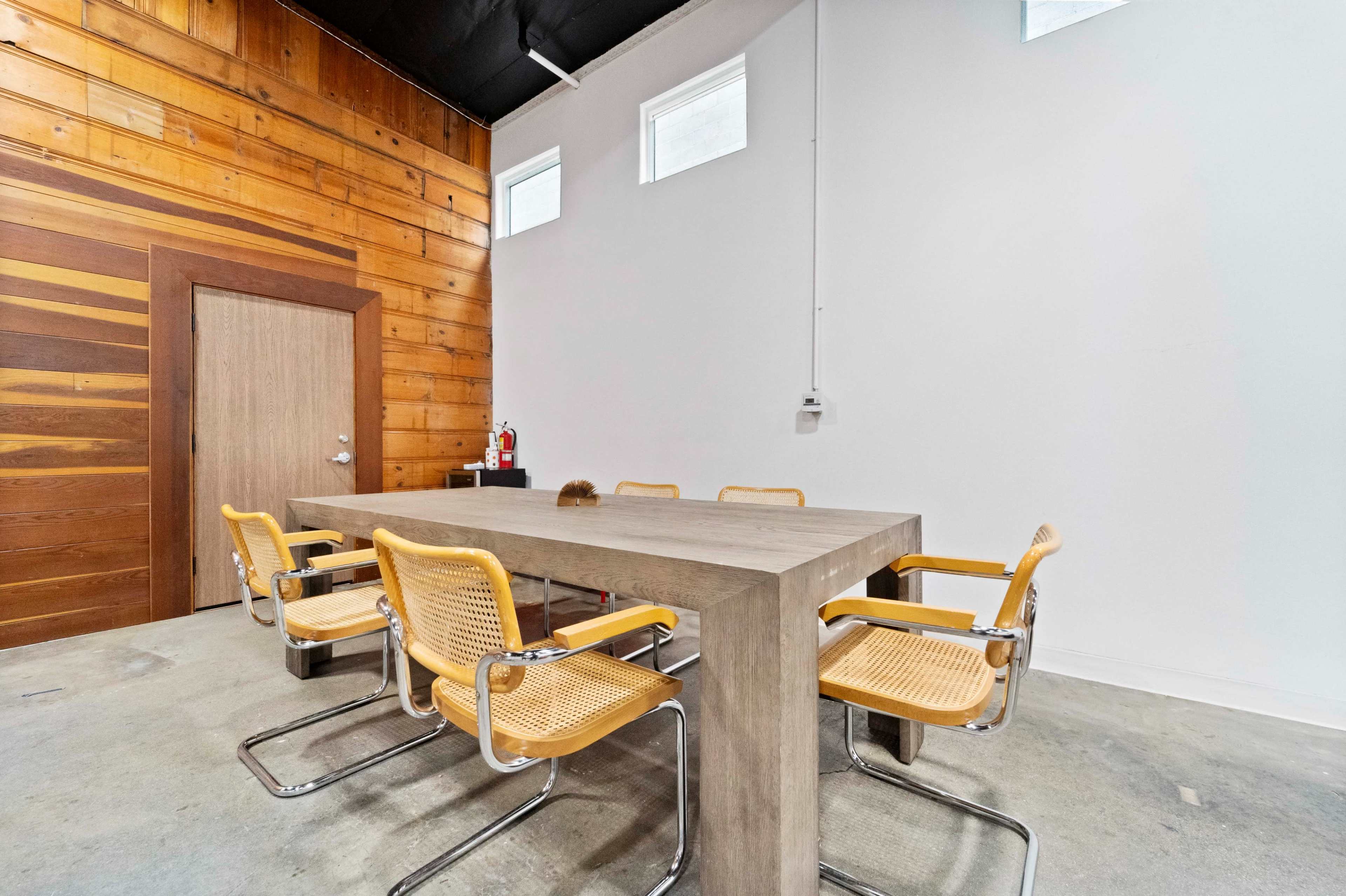 A meeting room features a long concrete table surrounded by five beige chairs against a backdrop of wooden walls and a plain white wall.