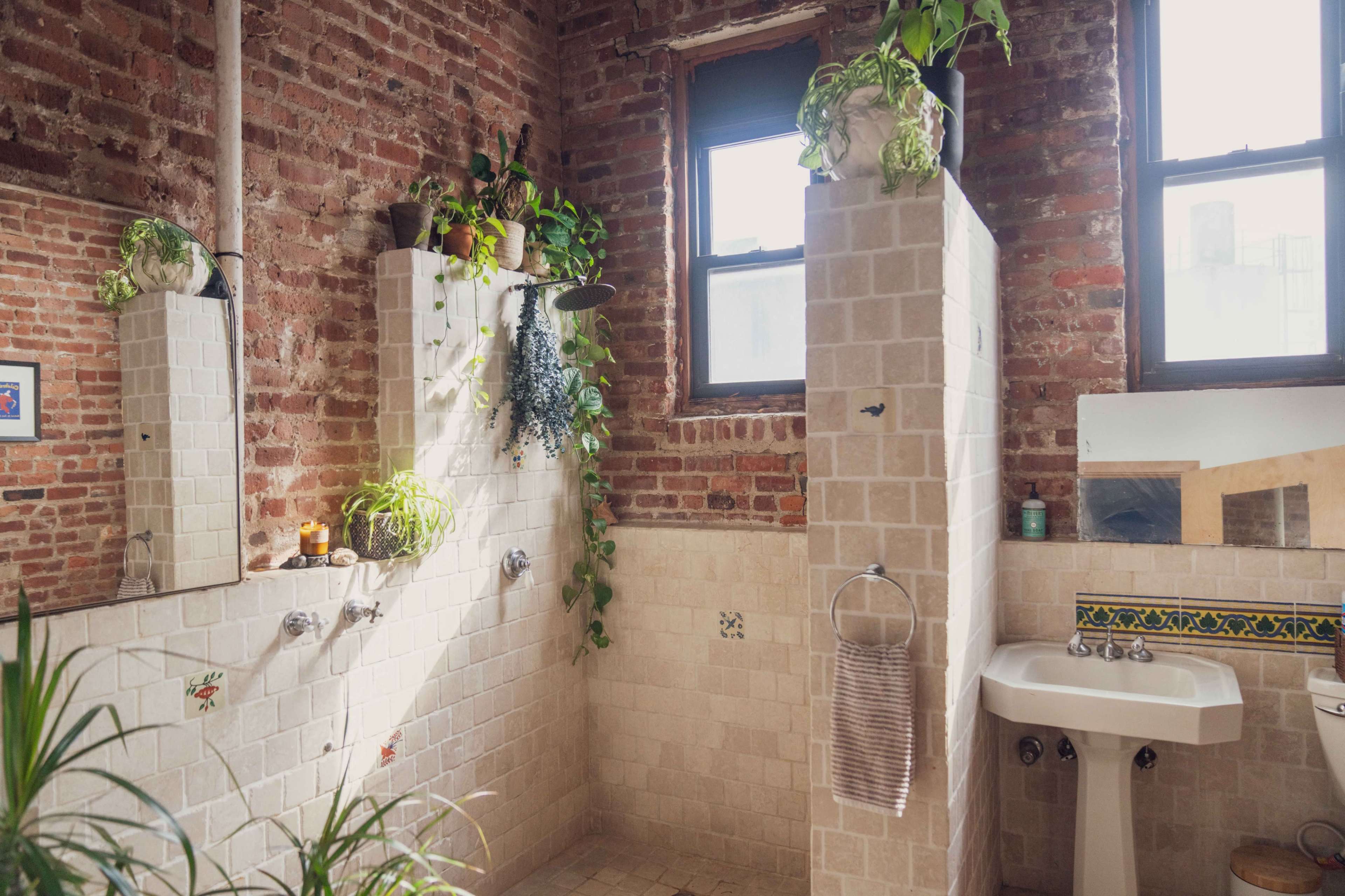 The image shows a brick-walled bathroom featuring two shower areas with tiled partitions, plants adorning the walls, and a sink near a window.