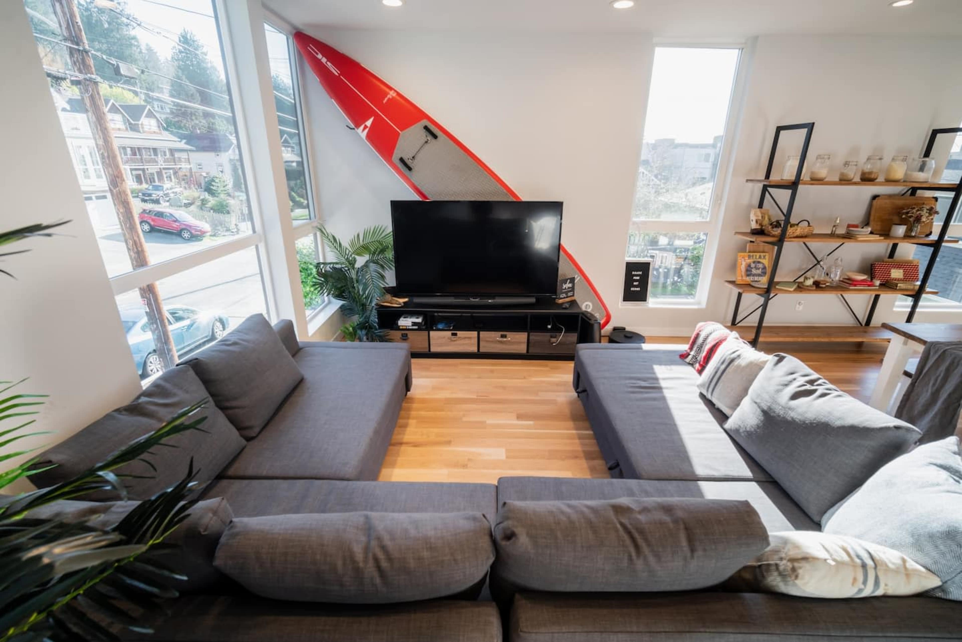 A modern living room features a large sectional sofa arranged in a U-shape around a television, with a red surfboard mounted on the wall as a decorative element.
