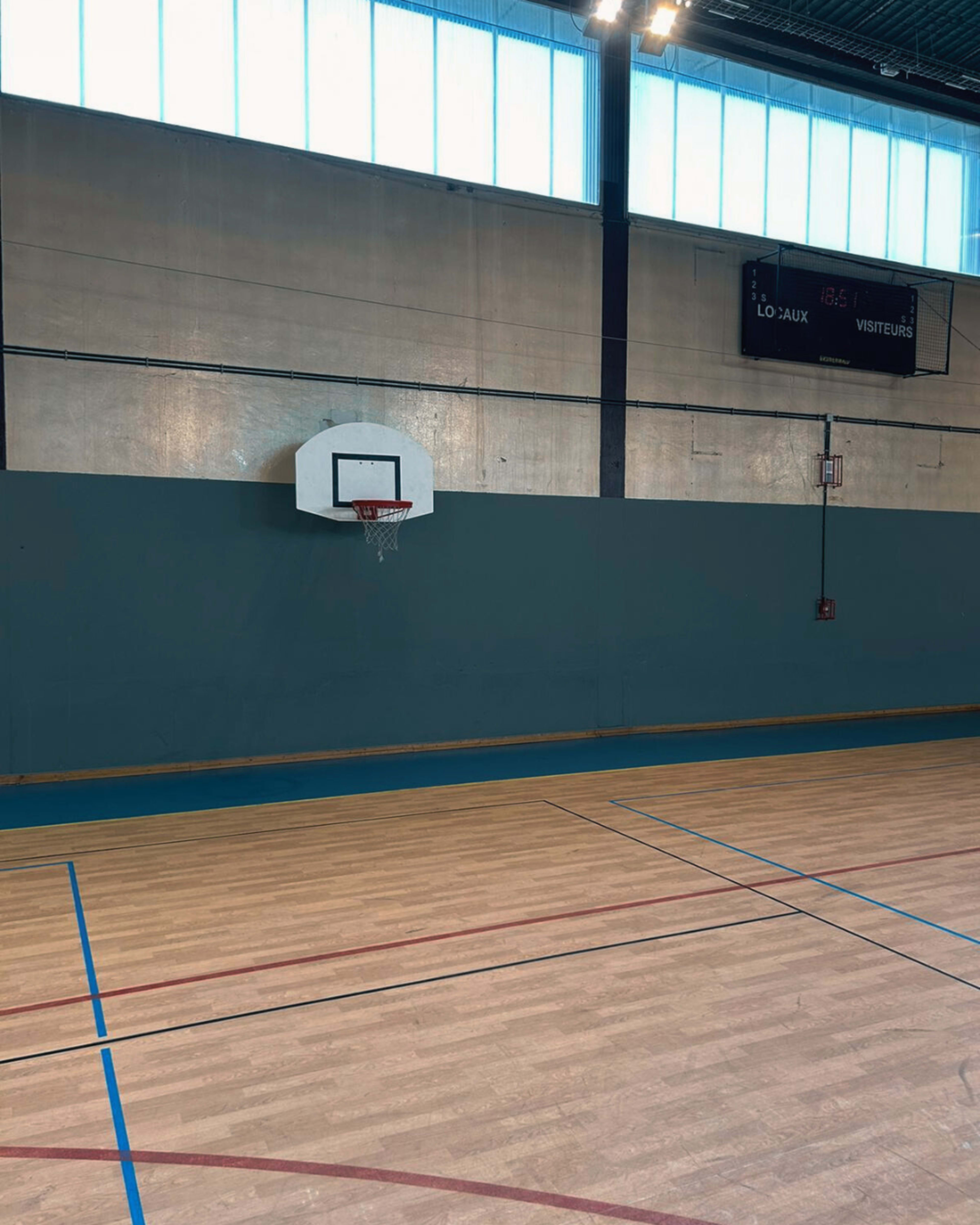A basketball hoop mounted on a wall inside a gymnasium with a wooden floor and blue markings.