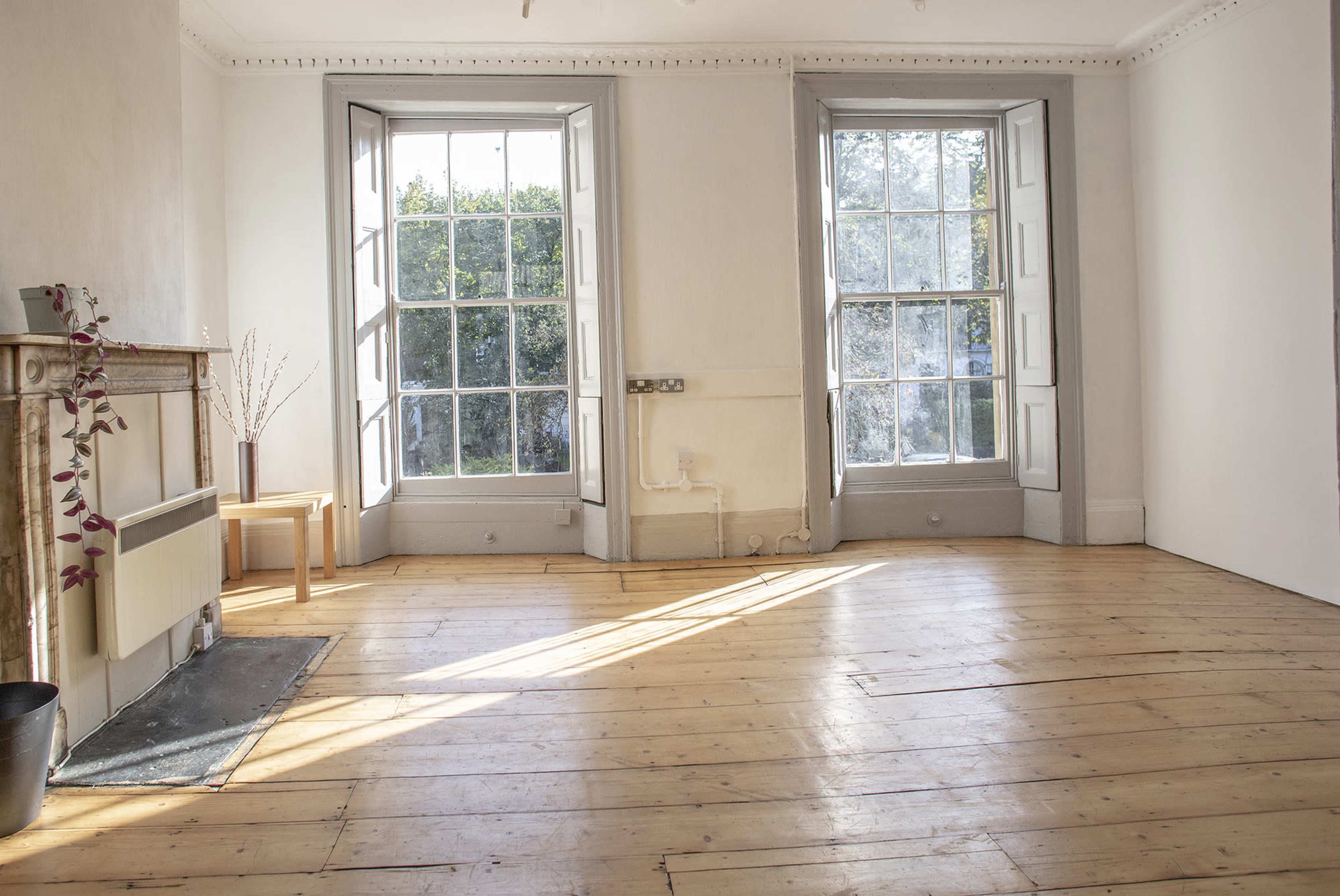 A room with two large windows on opposite walls, wooden flooring, and minimal furnishings including a small table and a decorative plant.