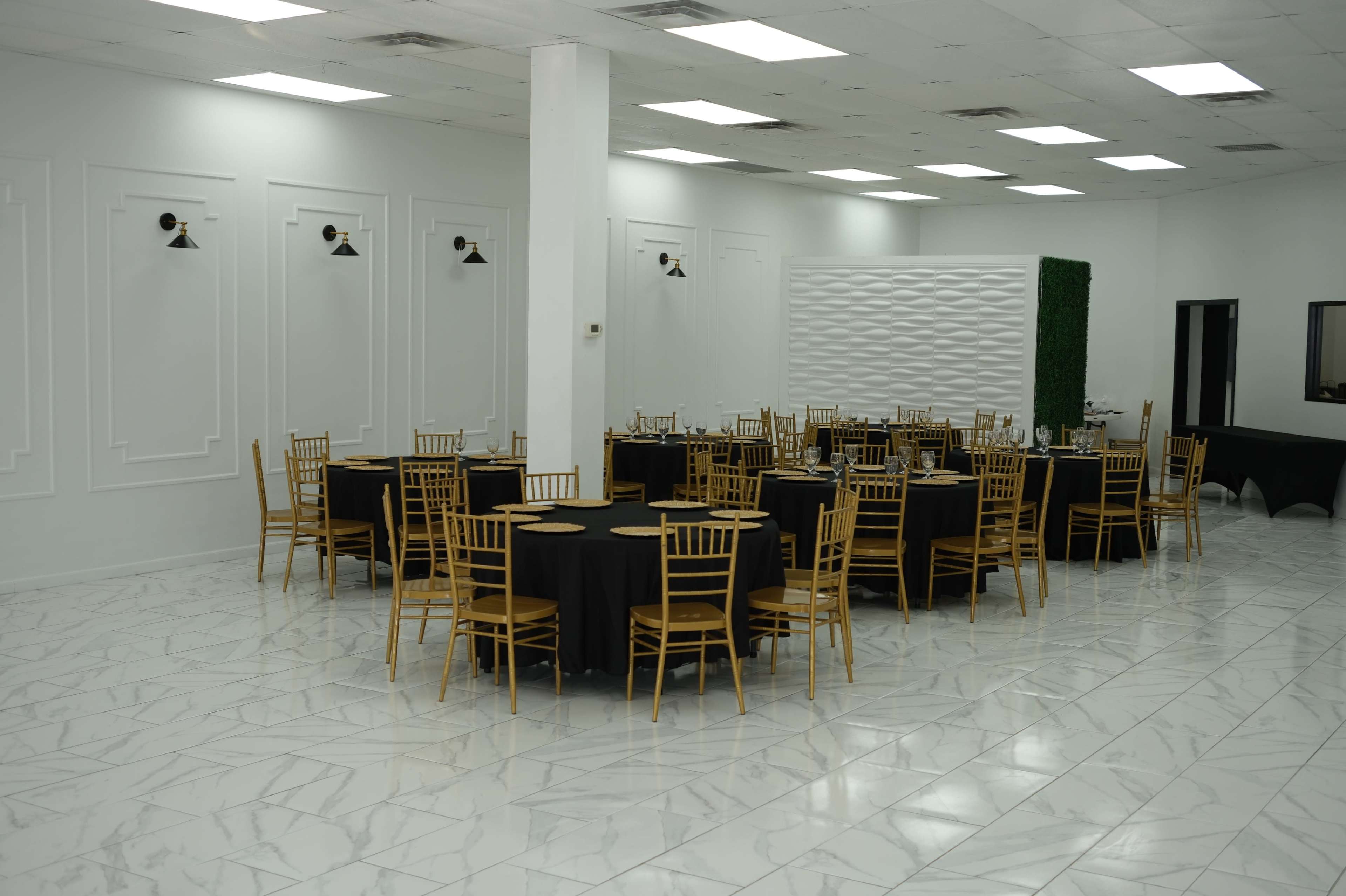 The image shows a spacious event hall featuring gold chiavari chairs arranged around black tablecloths on round tables, set against a white wall with decorative elements.