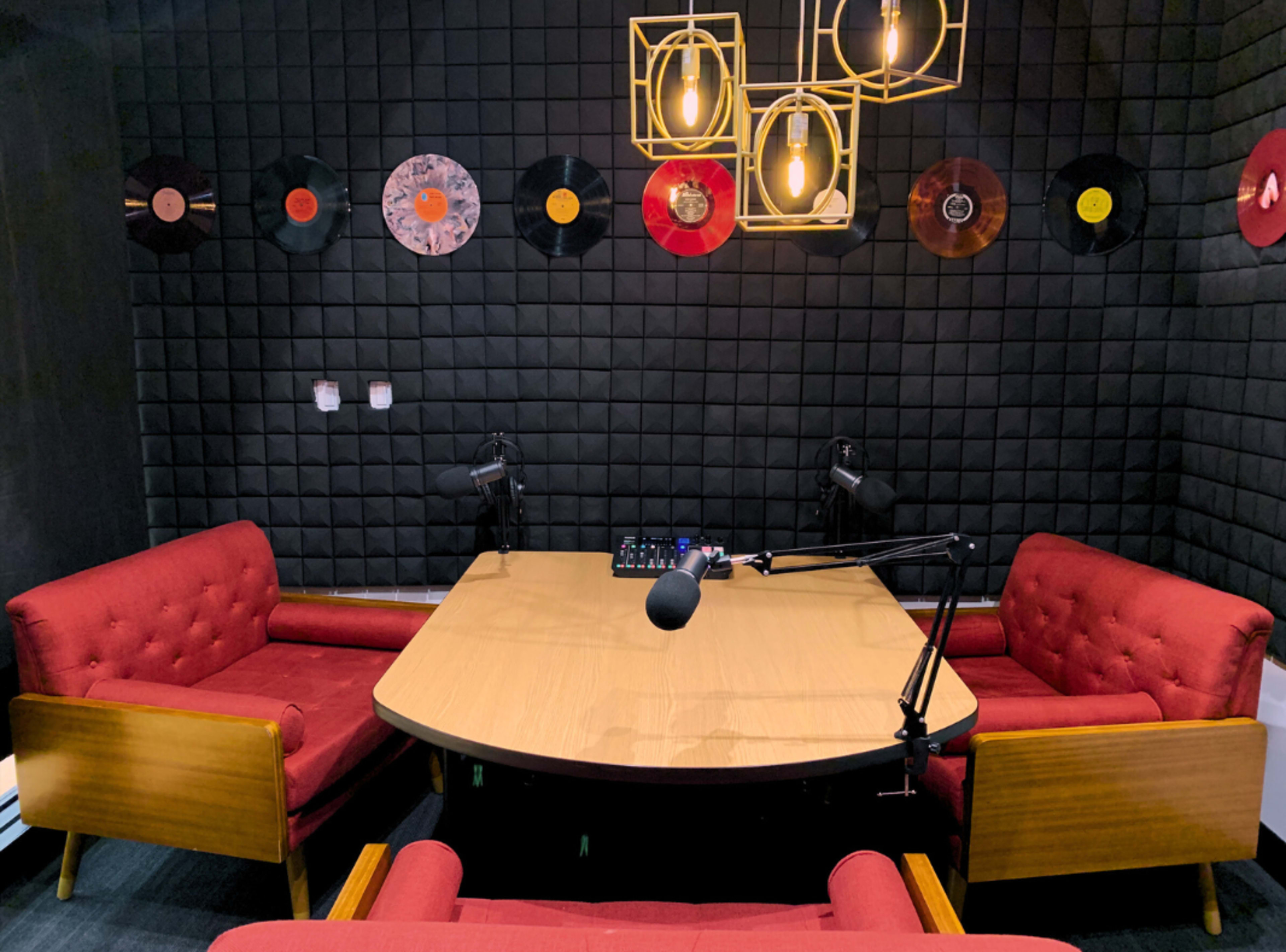 The image depicts a recording studio with a round wooden table, two red upholstered chairs, and several vinyl records displayed on a black wall.