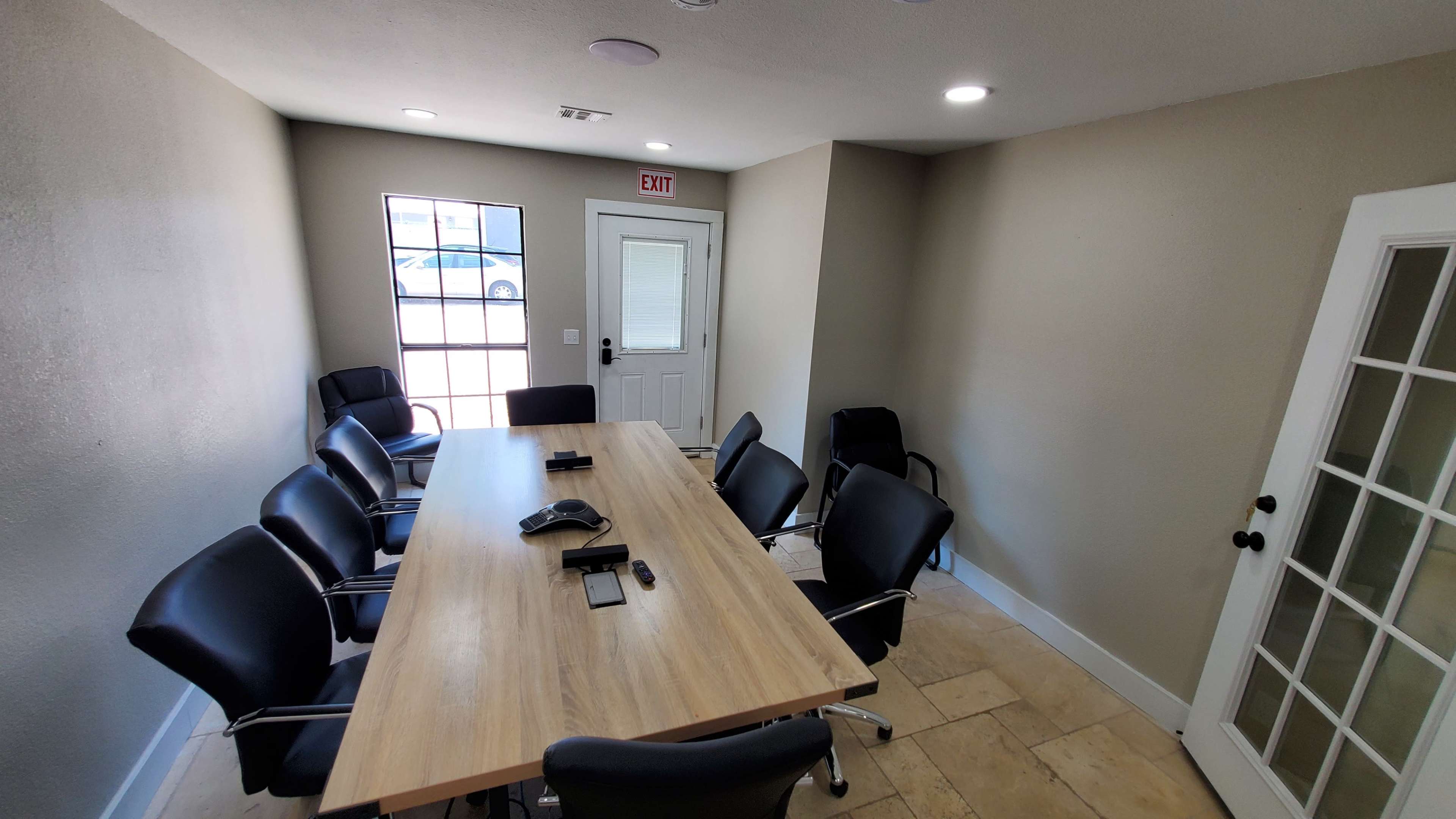 A conference room features a large wooden table surrounded by black chairs and a wall with a windowed door leading outside.