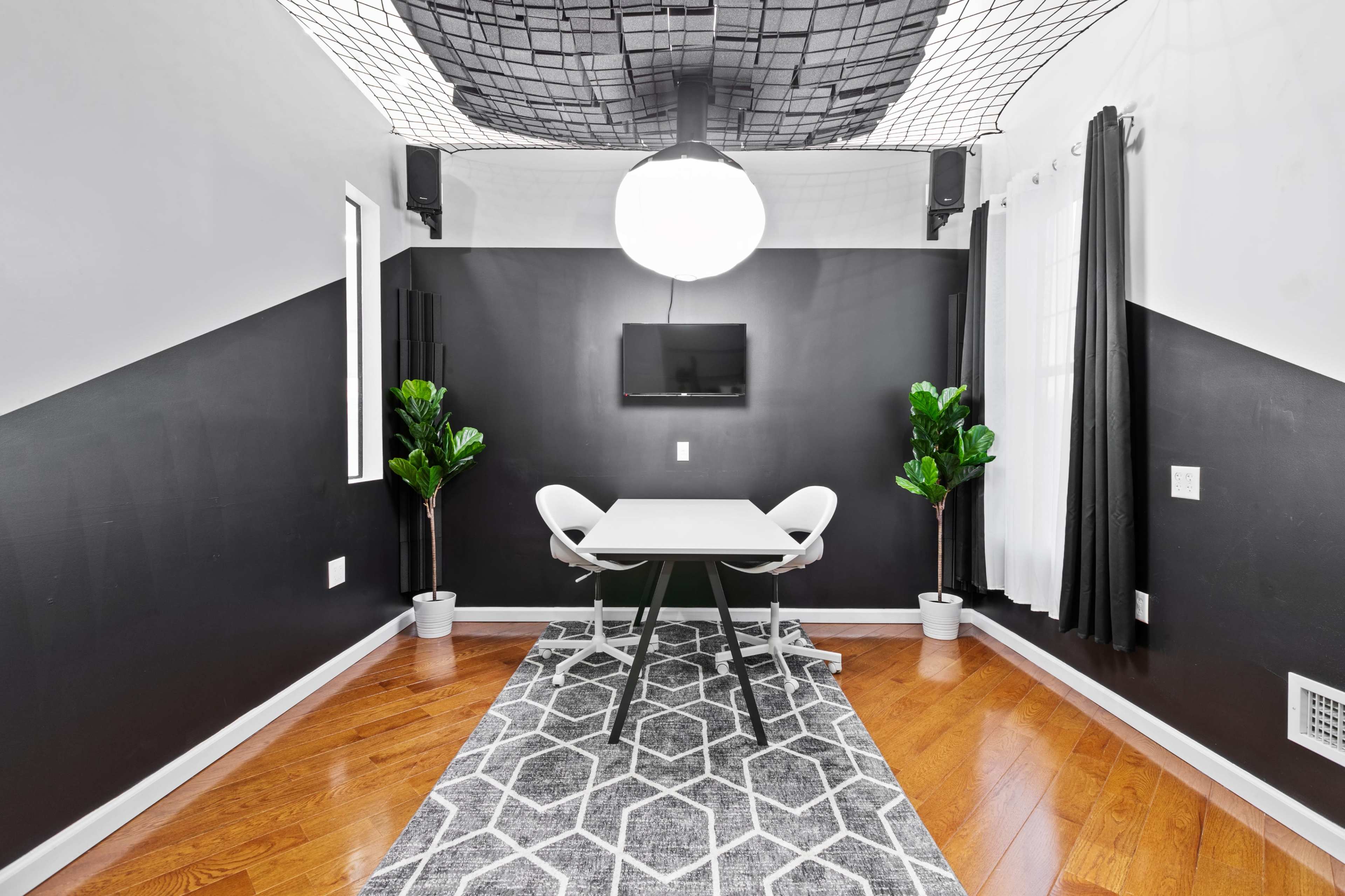 A small, modern meeting room features a table with two chairs, a large globe light fixture, and two potted plants on either side, set against black and white walls.
