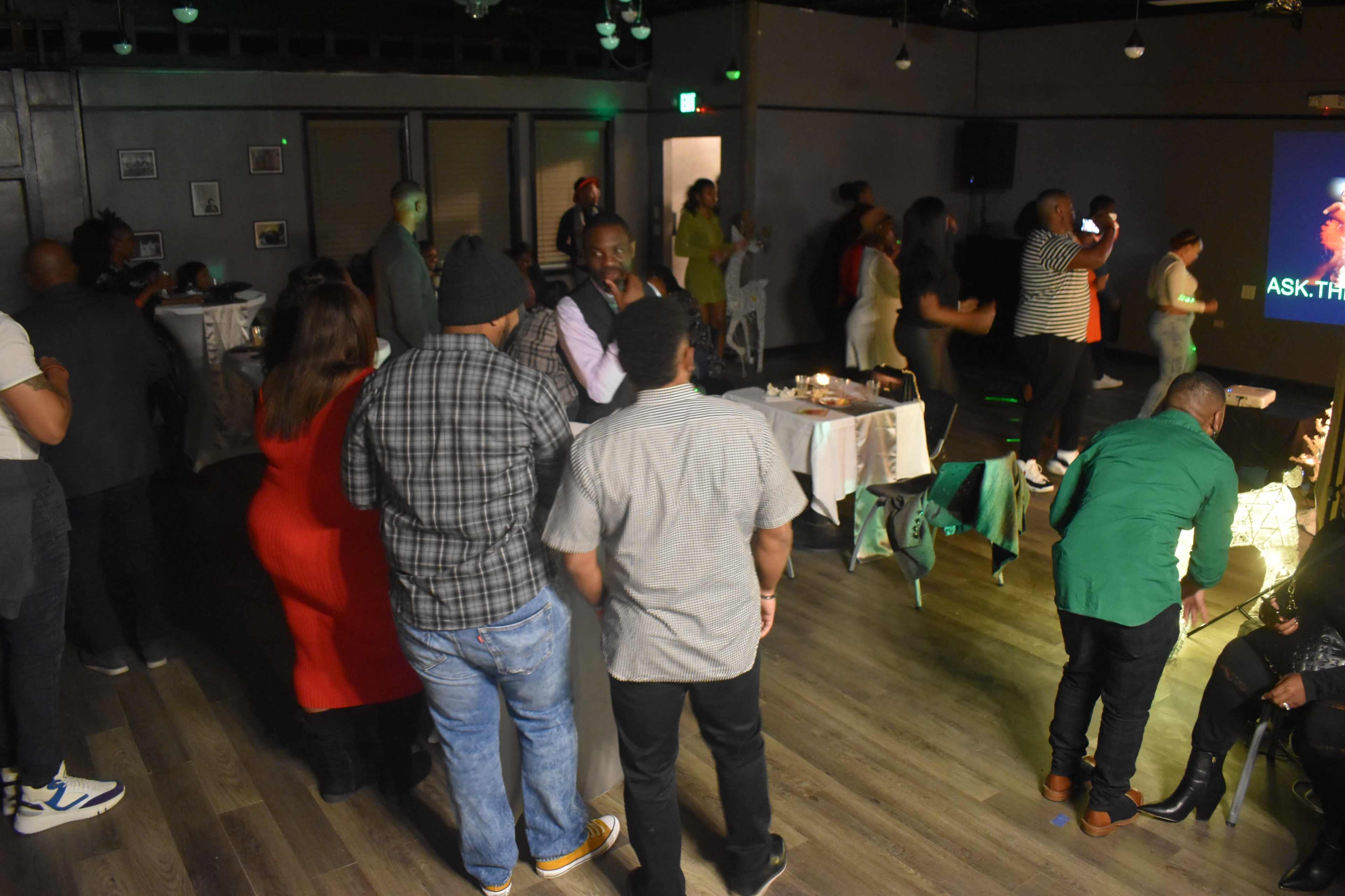 A group of people socialize in a dimly lit venue with tables and a projection screen displaying text, while some attendees dance in the background.