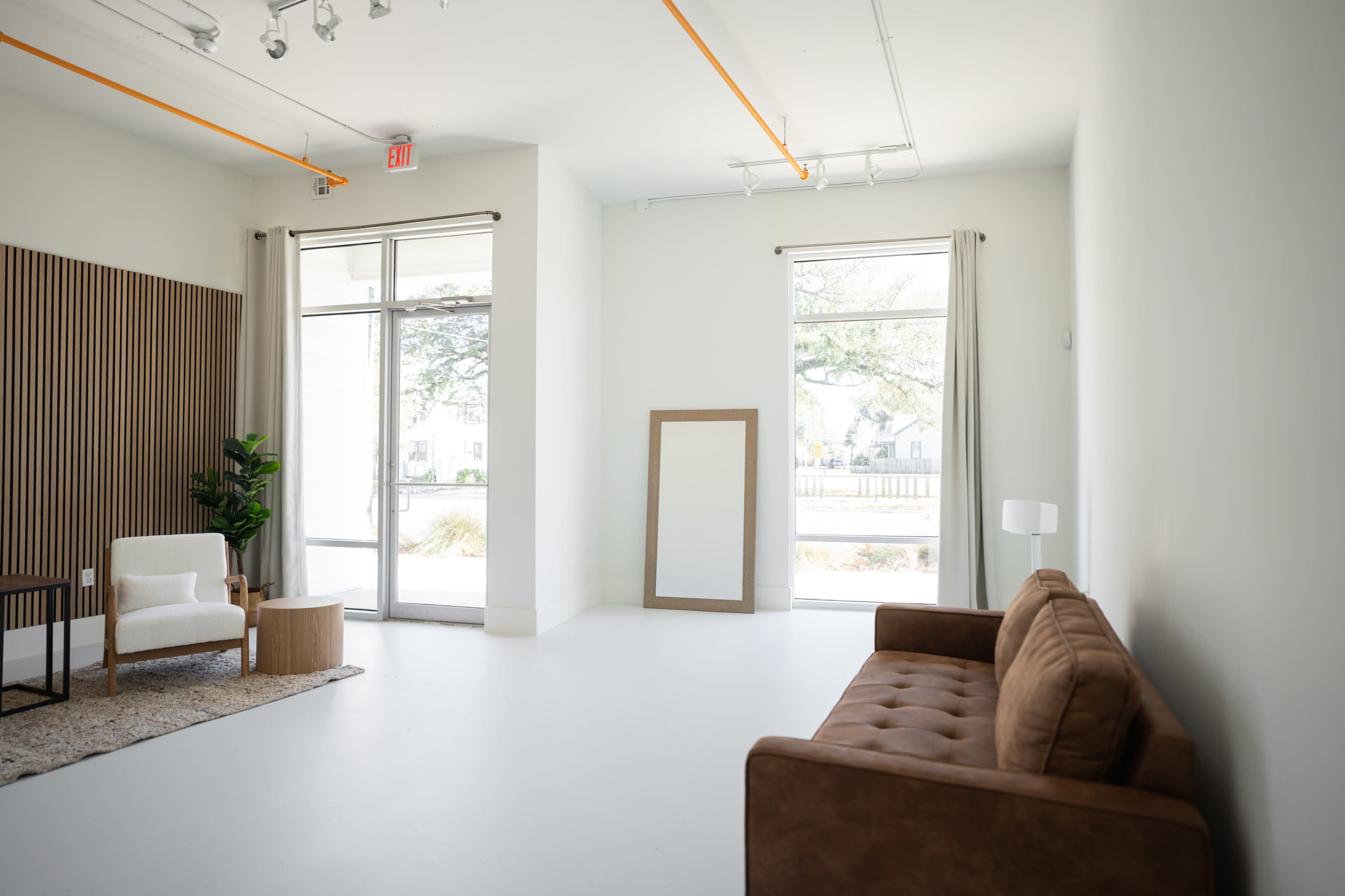 A minimalist interior space featuring a brown sofa, a small wooden table, a large mirror, and large windows with natural light.