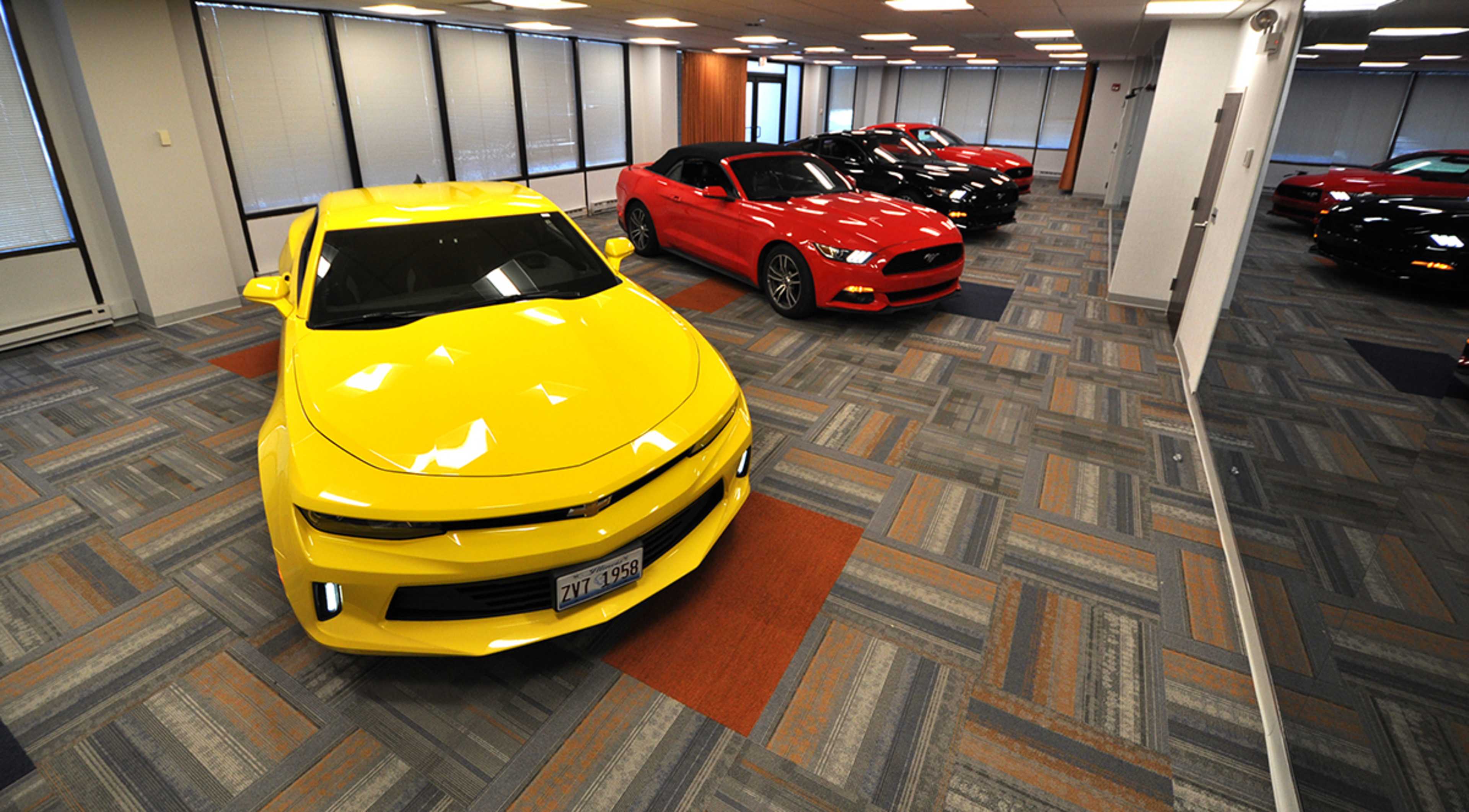 A bright yellow car is parked in the foreground of a showroom featuring several other cars, including a red convertible and black vehicles, arranged in a well-lit space with patterned flooring.