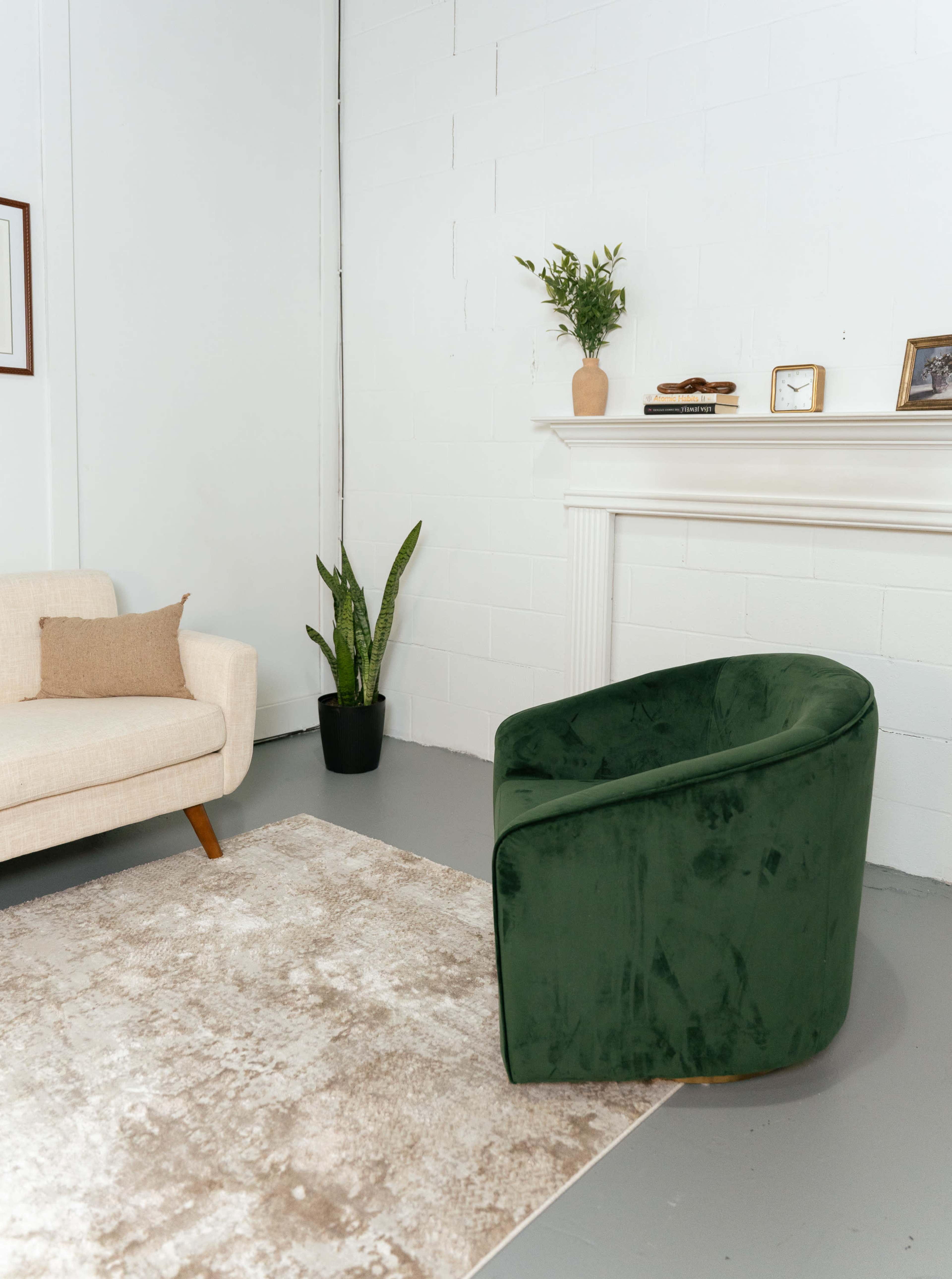 A green curved chair and a light-colored sofa are positioned in a minimalist room with a white wall and a decorative mantelpiece.