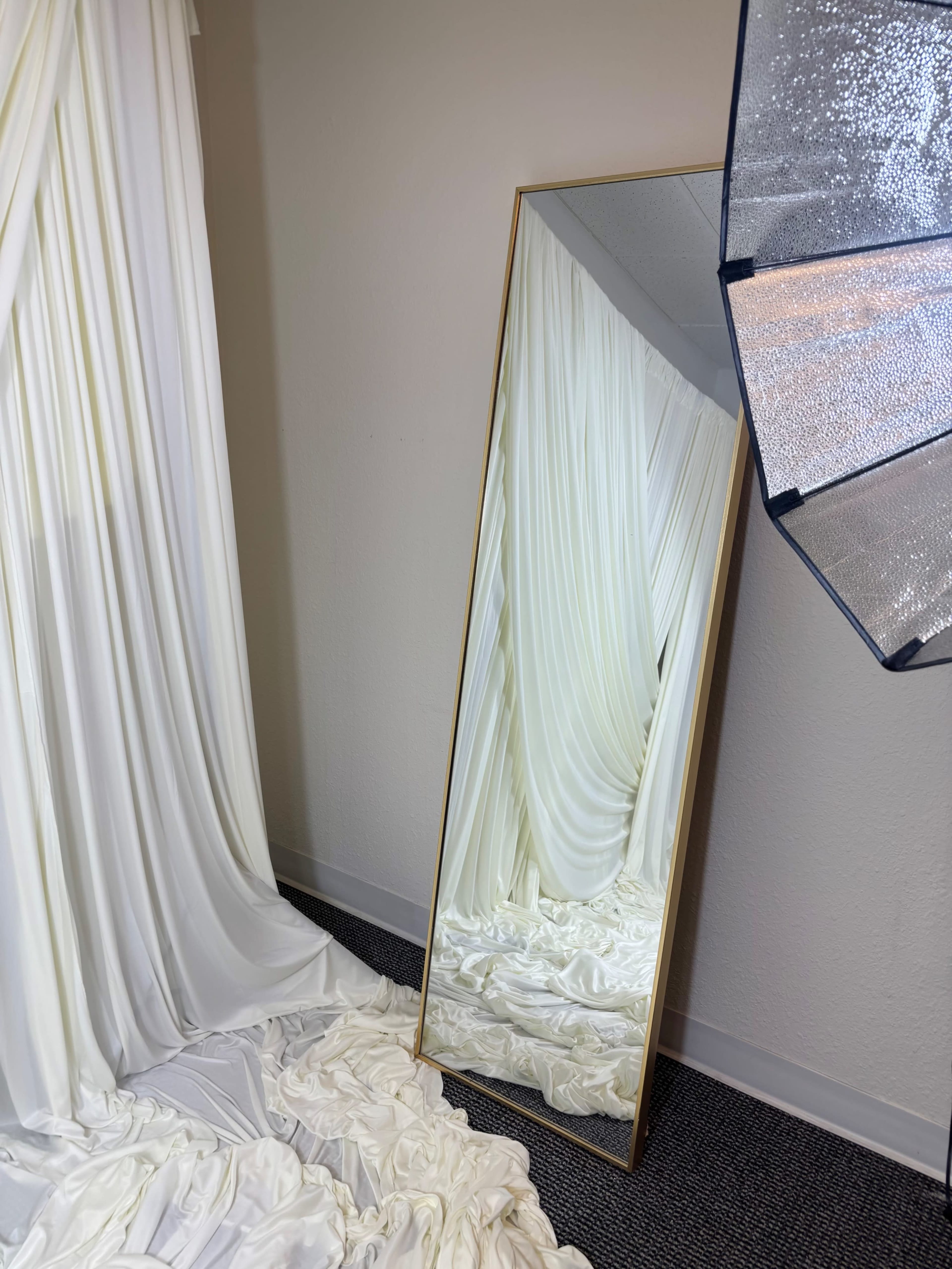 A large, framed mirror stands against a wall, reflecting draped white fabric and a softly illuminated area on the floor.