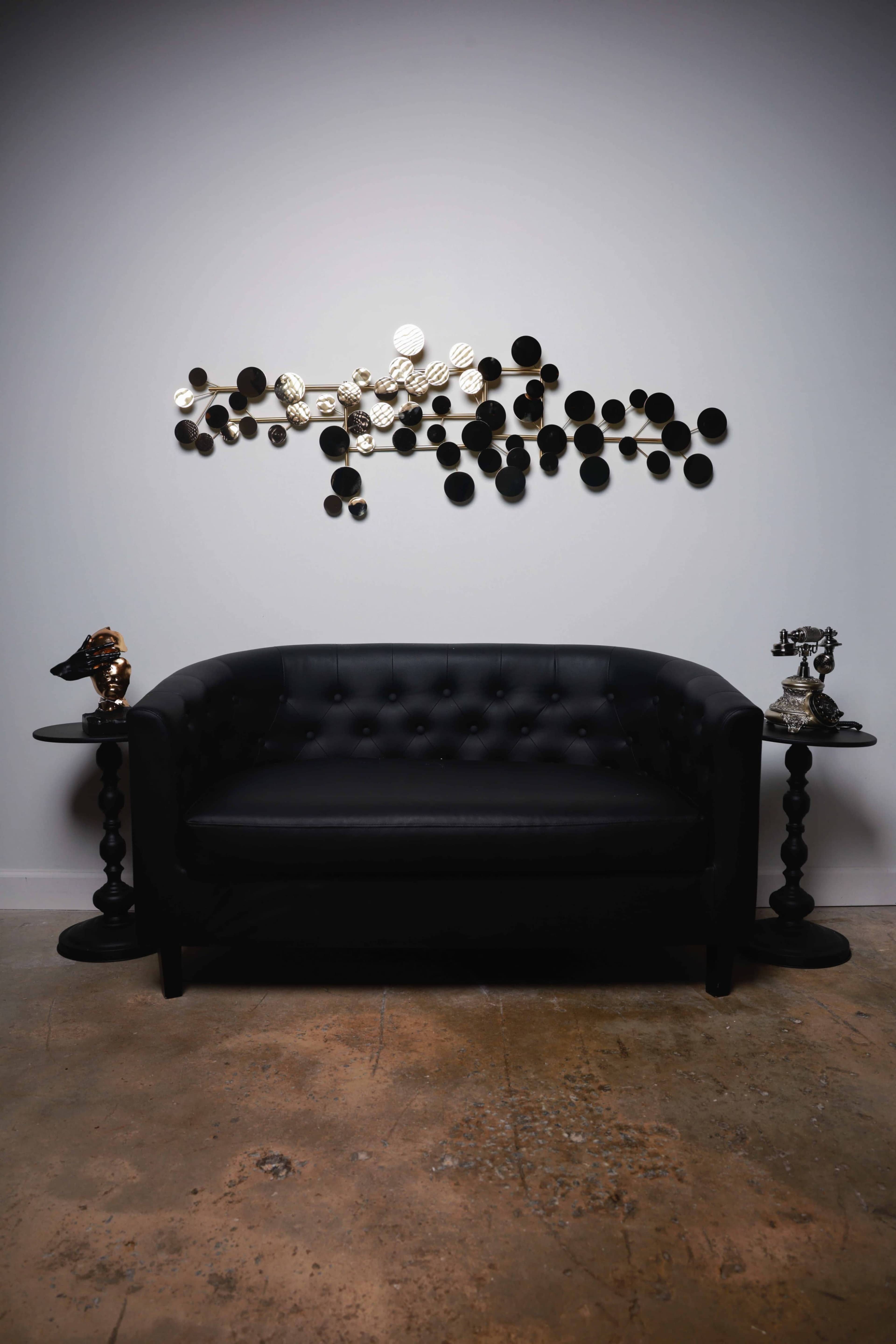 A black tufted sofa is positioned between two round side tables, with a decorative wall piece of circular shapes hanging above.