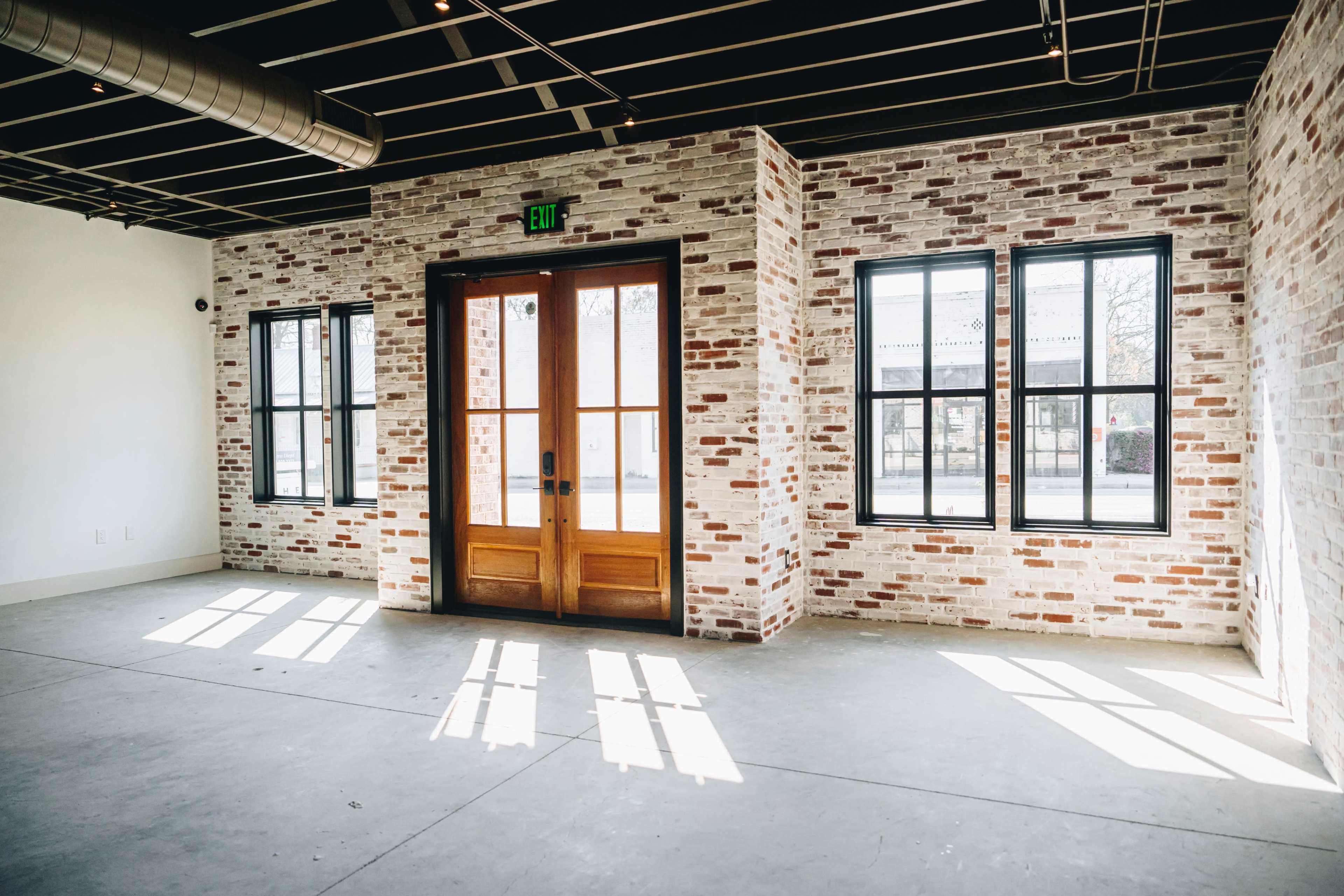 A room with brick walls, large windows, and a double wooden door, featuring sunlight casting shadows on the concrete floor.
