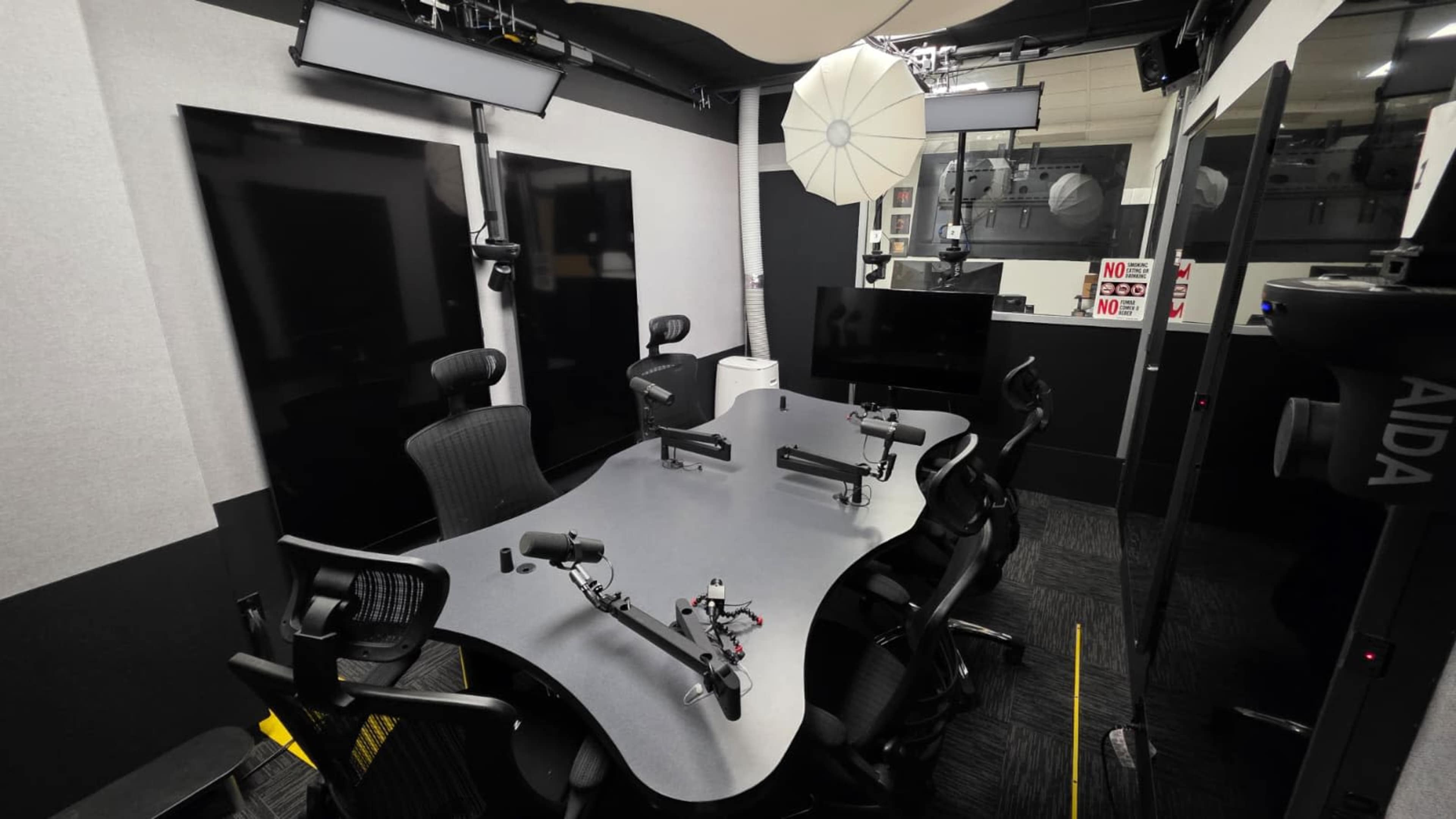 The image shows a modern conference room equipped with a large oval table, multiple chairs, and various microphones, alongside screens mounted on the walls.