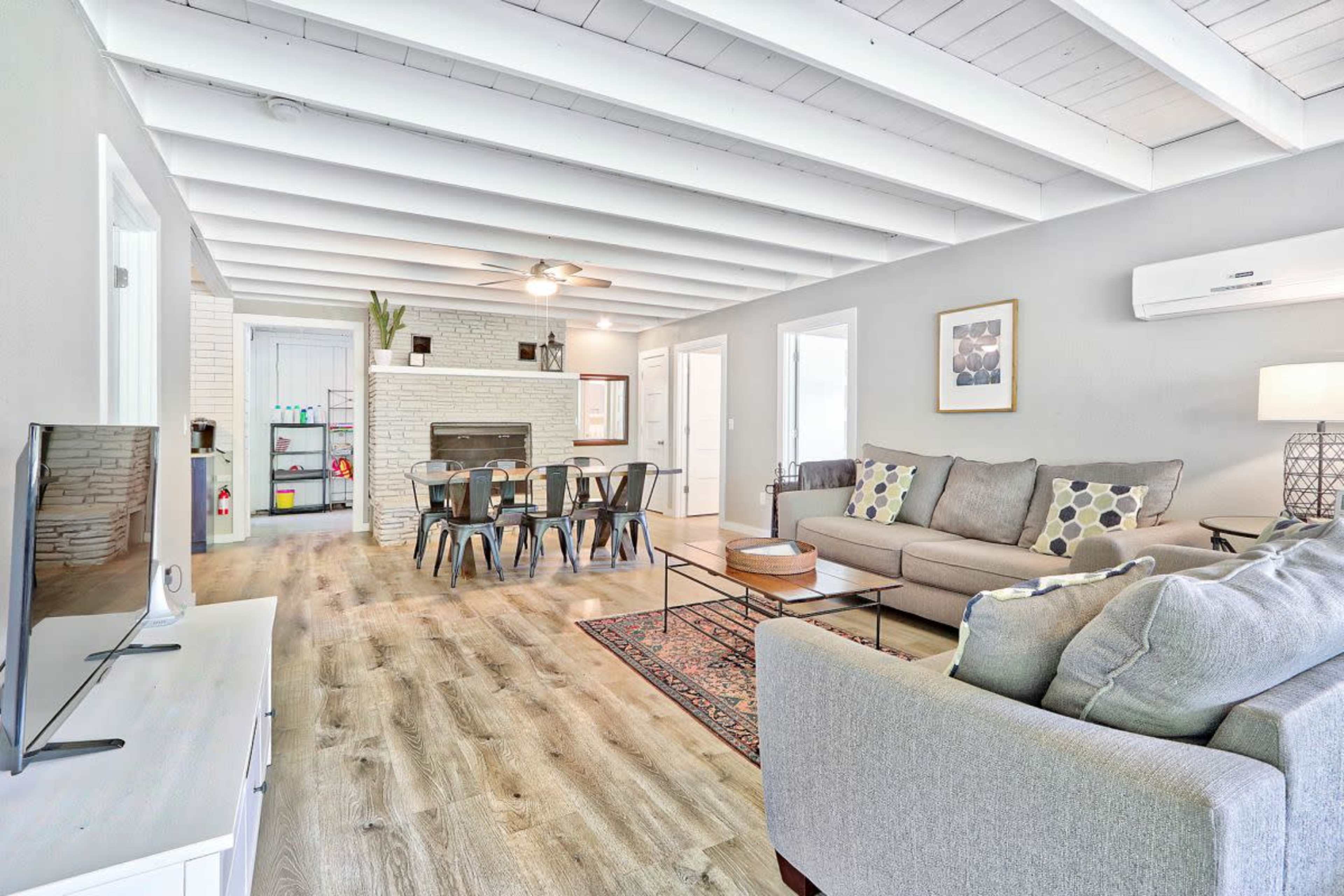 A modern living room features a seating area with a sofa, a coffee table, and a dining space with a long table, illuminated by ceiling fans and natural light from windows.