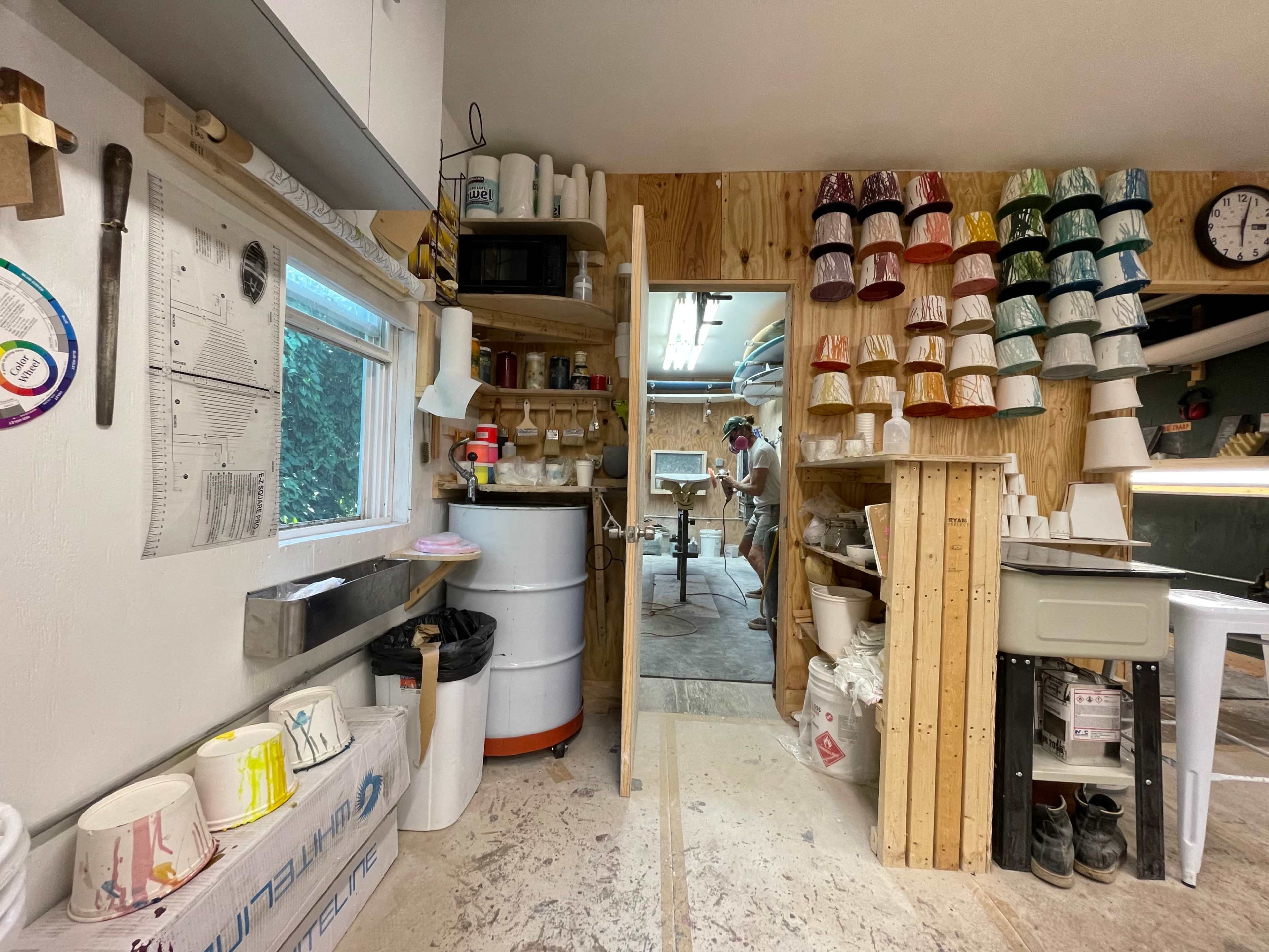 The image shows an organized workshop with shelves holding colorful paint cans and various tools, leading into a workspace area in the background.
