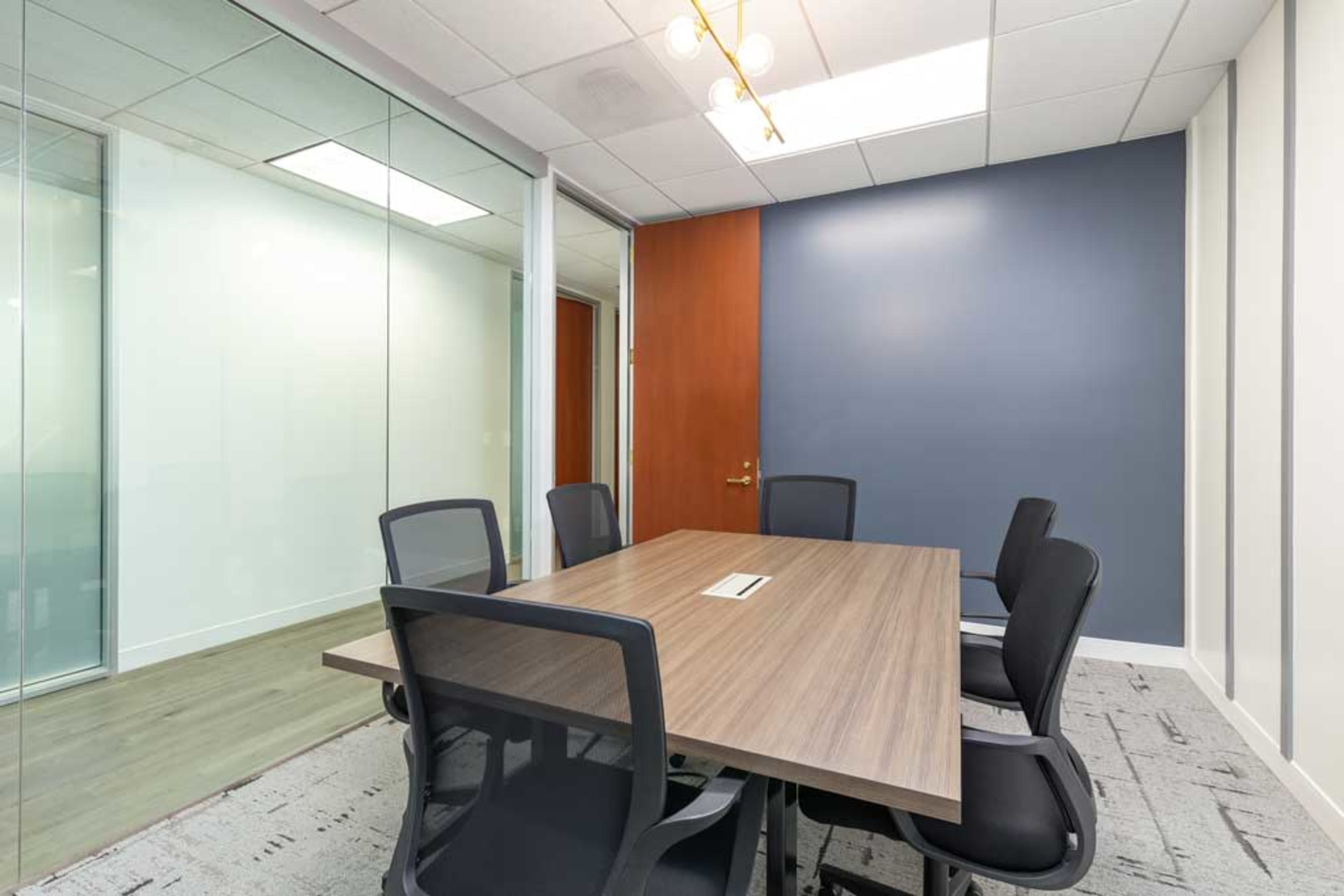 A conference room features a wooden table surrounded by six black chairs, with glass walls and a blue accent wall.