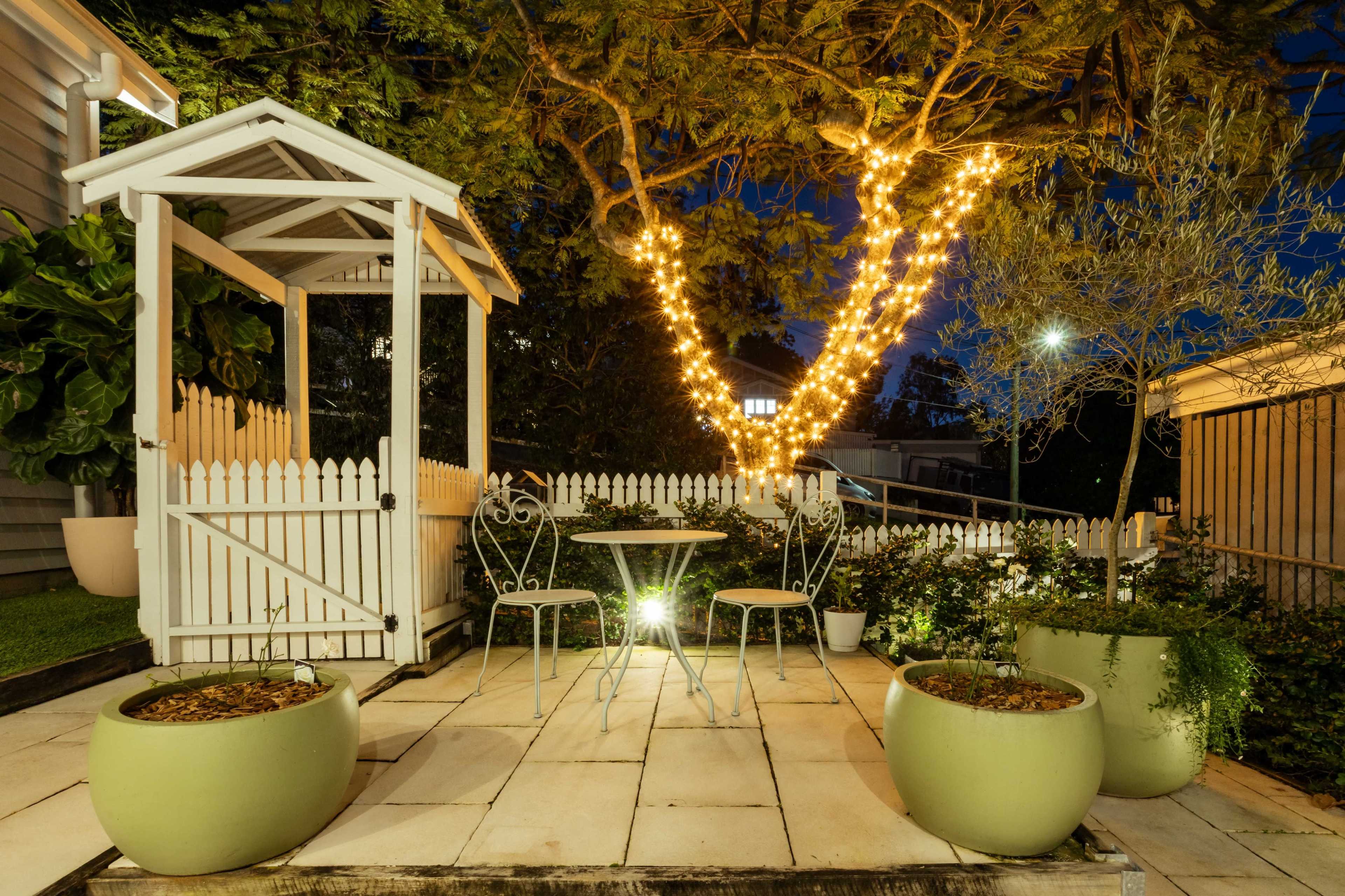 A cozy outdoor seating area features a white gazebo and a small table with chairs illuminated by string lights in the evening.