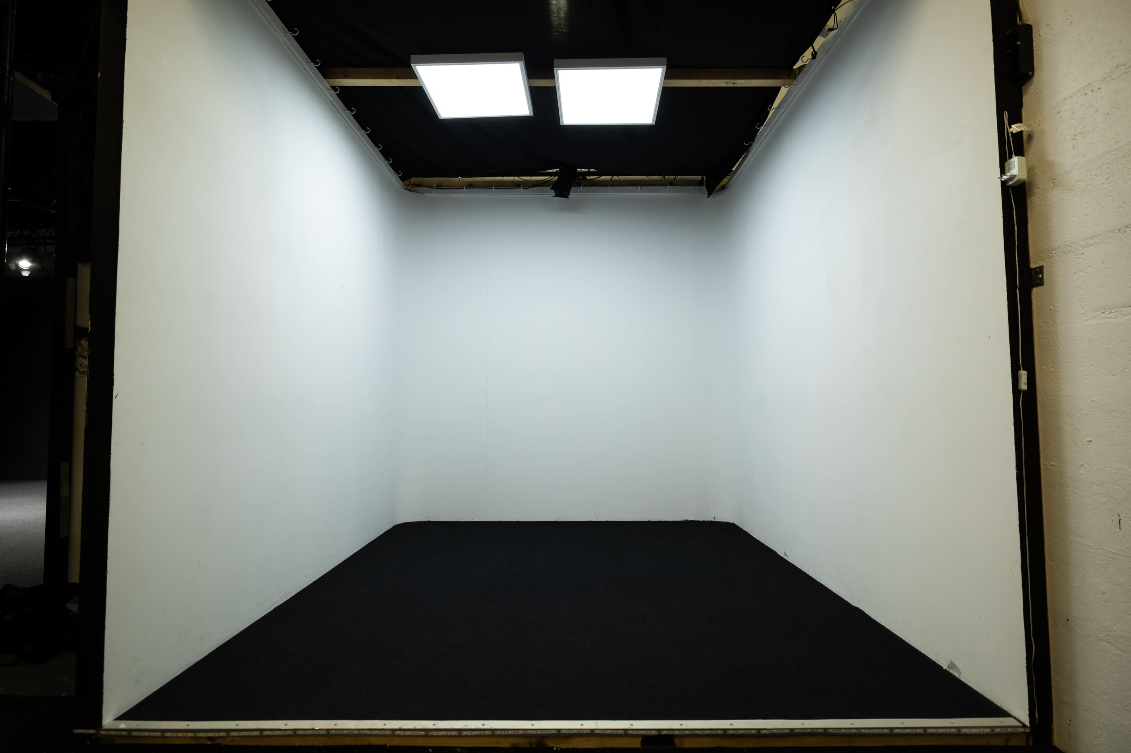 An empty room with white walls and a dark carpet, illuminated by two overhead lights.