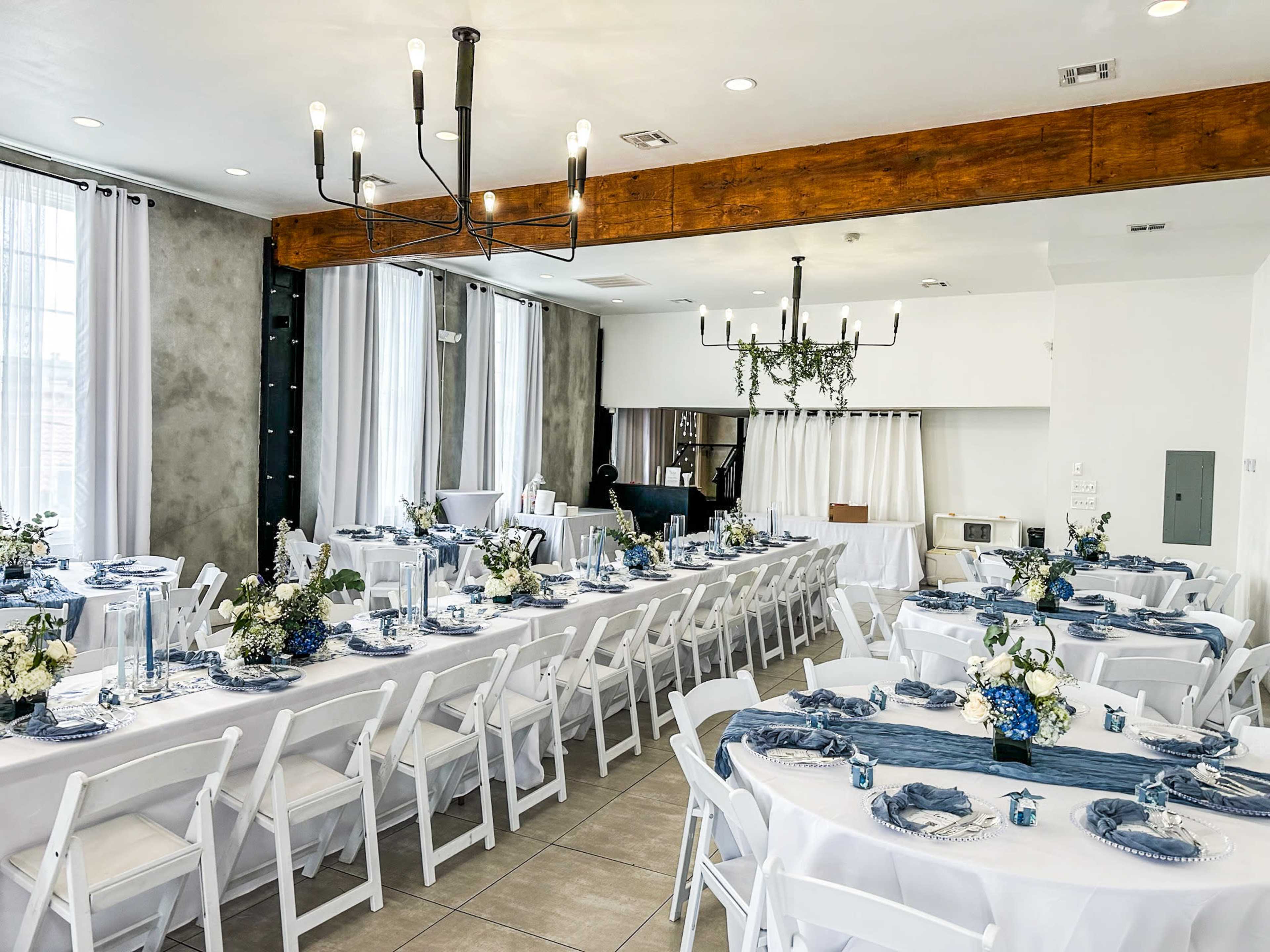 The image shows a neatly arranged dining space with tables set for an event, featuring white tablecloths, blue decorations, and floral centerpieces.