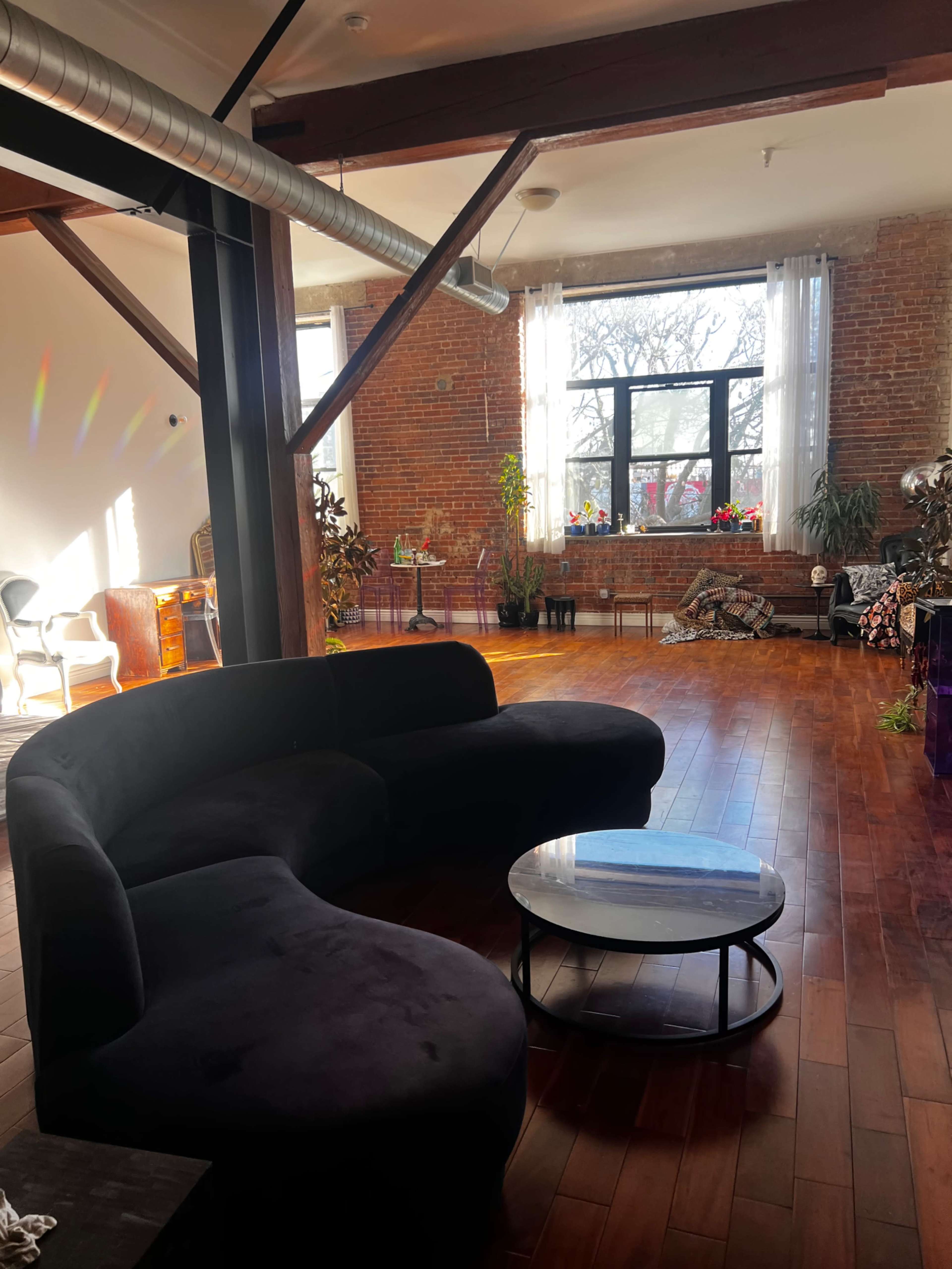 The image depicts a spacious, modern room with a curved black sofa and a round coffee table on wooden flooring, illuminated by natural light from large windows.