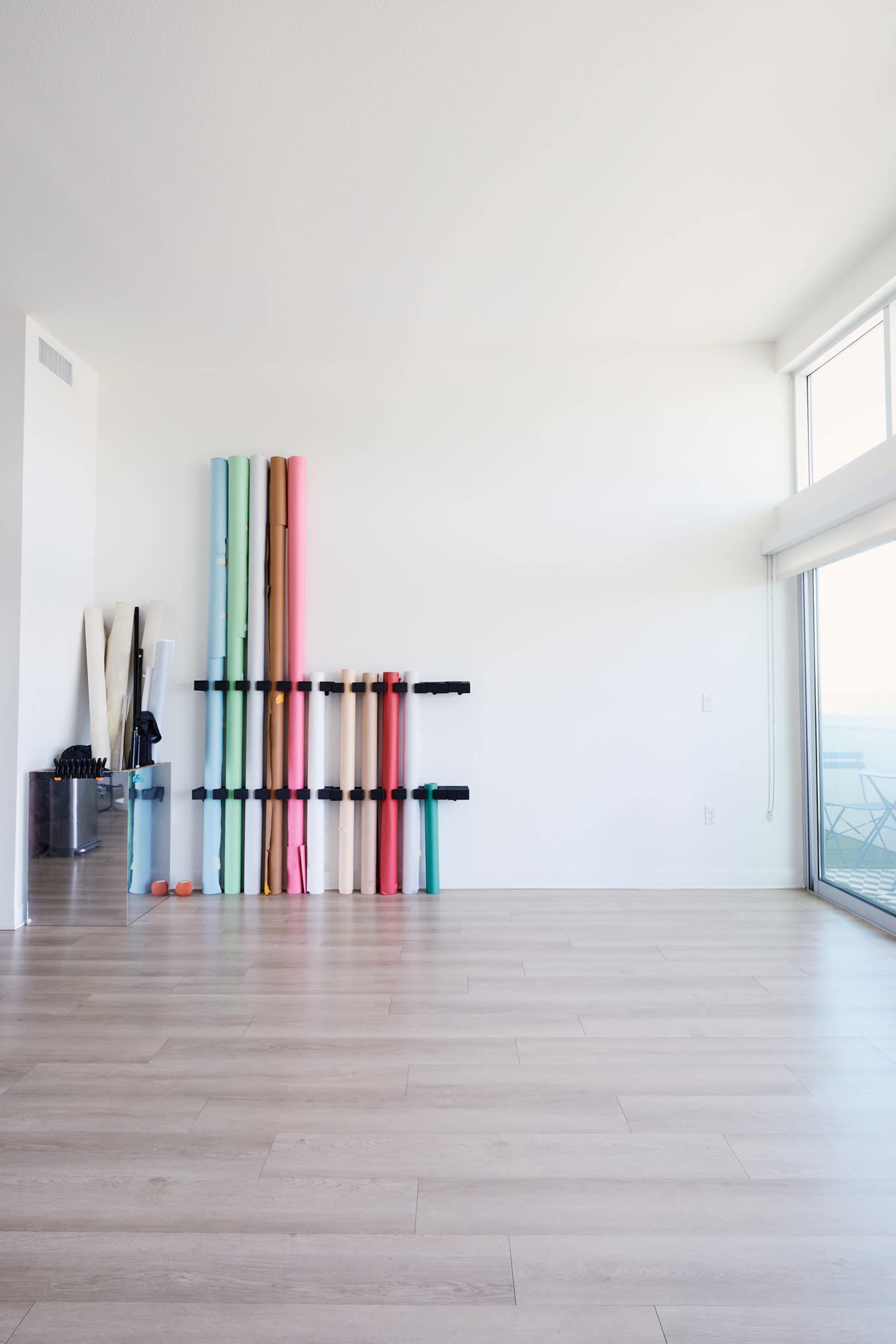The image shows a spacious, well-lit room with light wooden flooring and a wall-mounted storage rack holding various colored exercise mats.