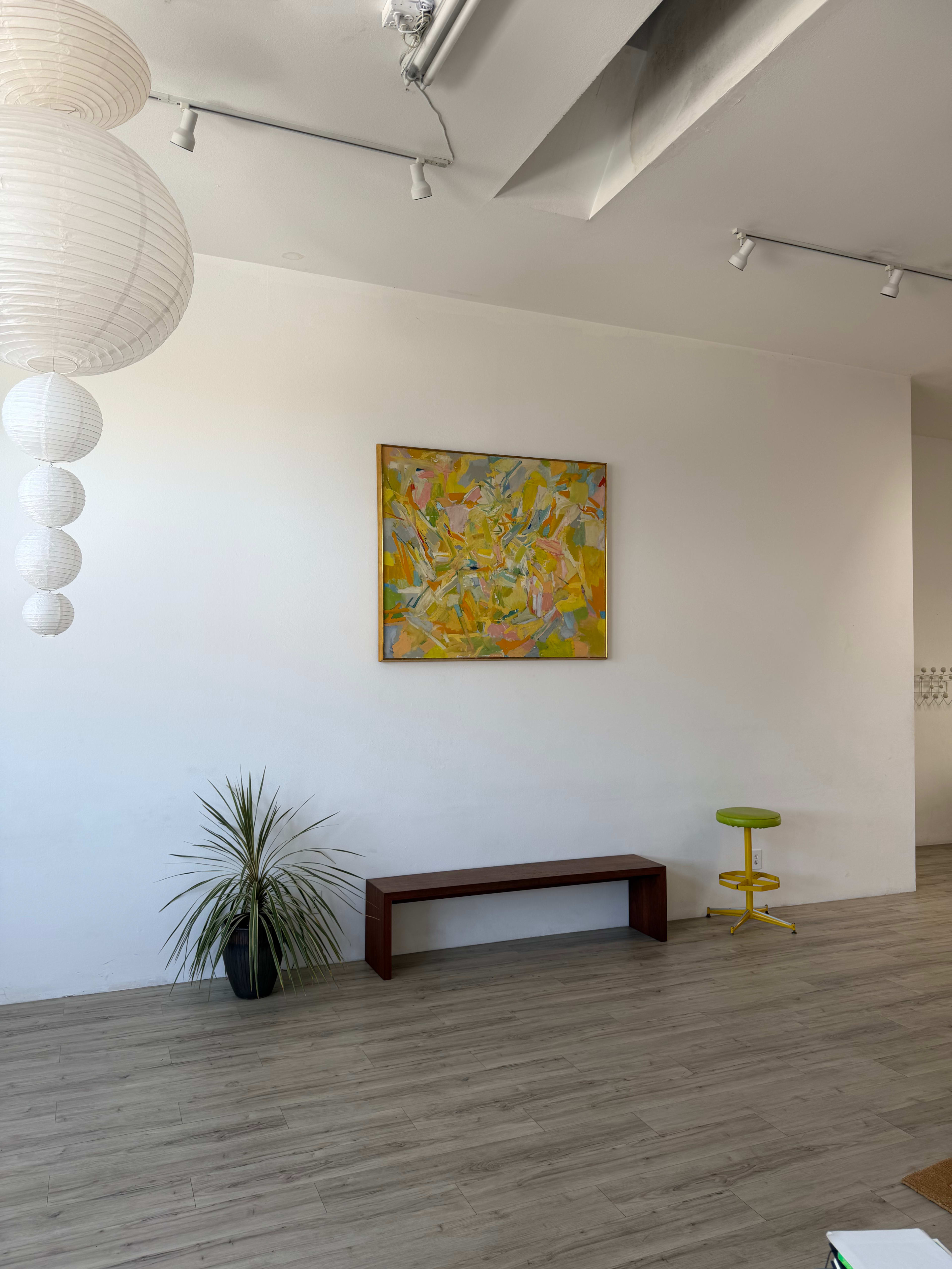 The image shows a modern, minimalist interior featuring a wooden bench, a plant, a colorful painting on the wall, and a yellow stool.