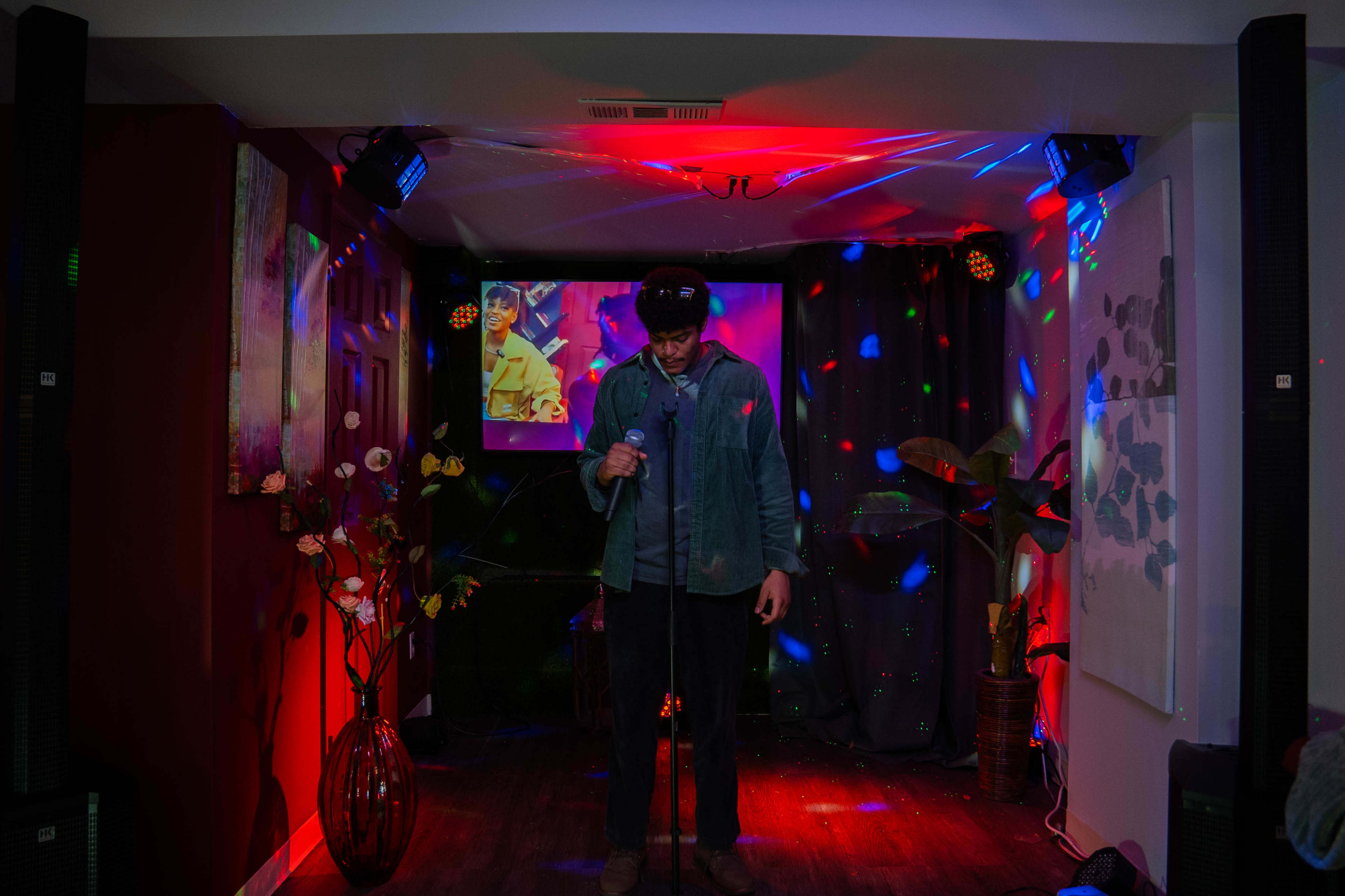A person stands on a stage with a microphone in a dimly lit room illuminated by colorful lights and a video screen in the background.