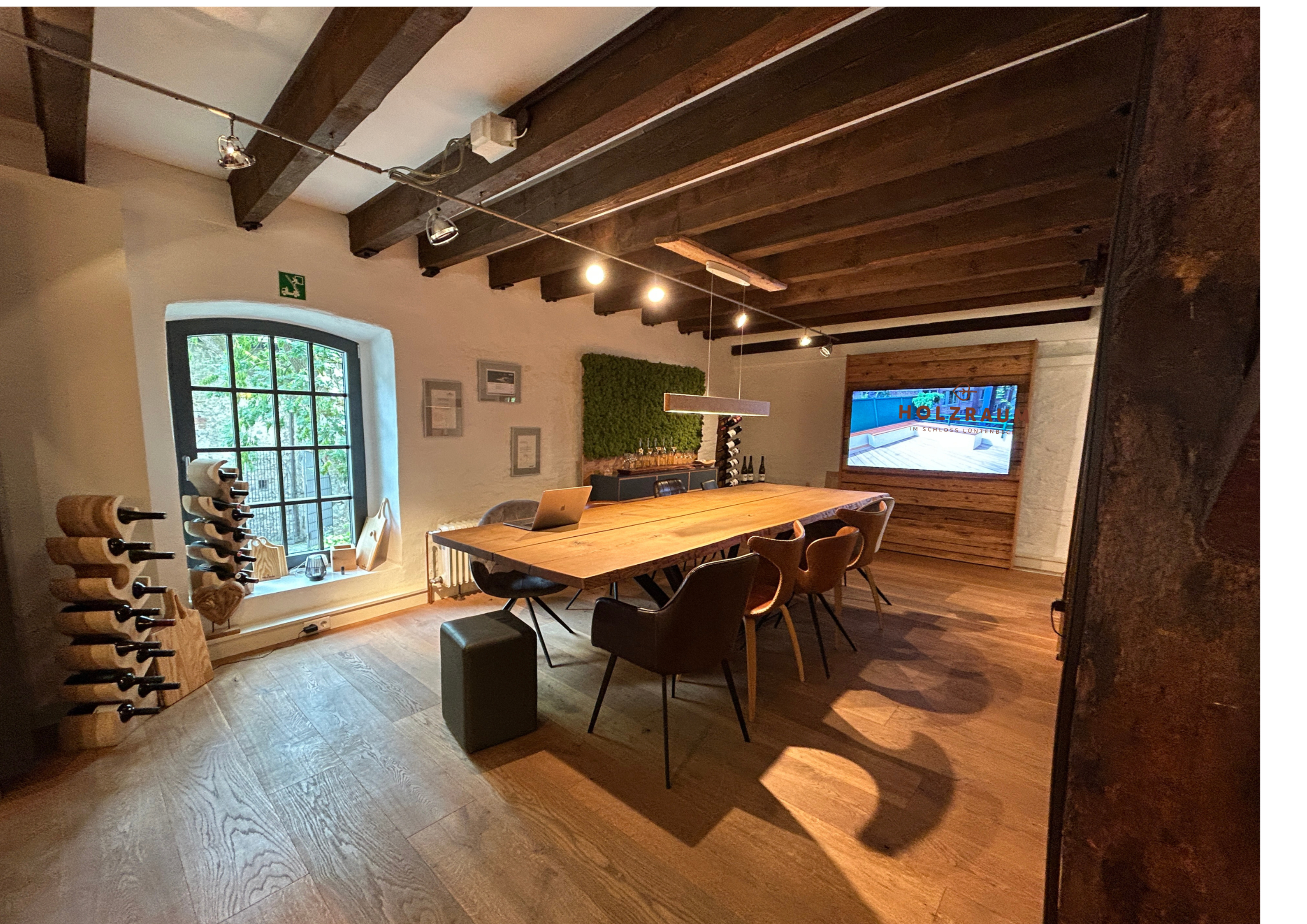A spacious meeting room features a large wooden table surrounded by chairs, a wall-mounted screen, and a wine rack beside a window.