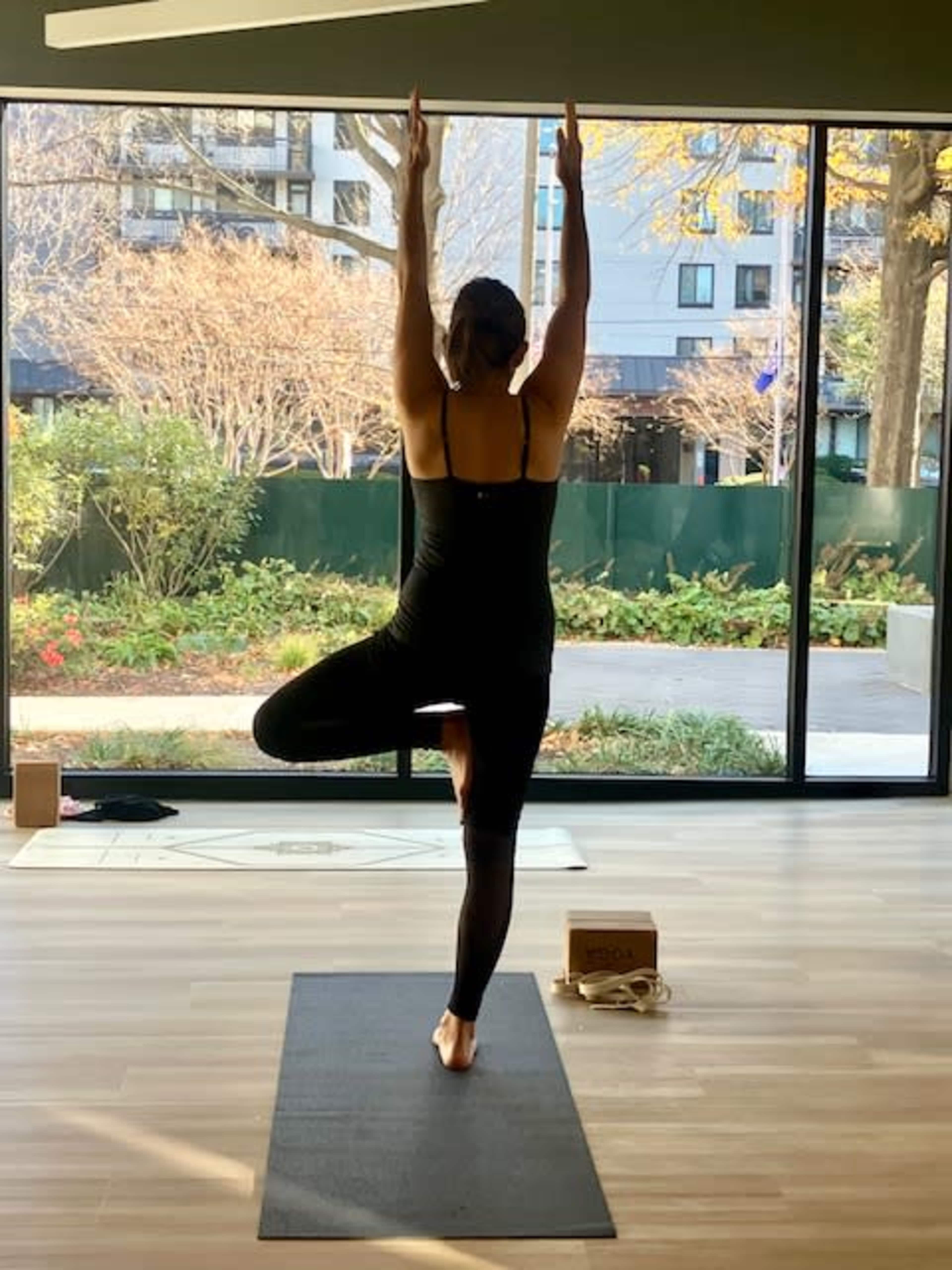 A person performs a yoga pose on a mat indoors, with large windows revealing a garden outside.