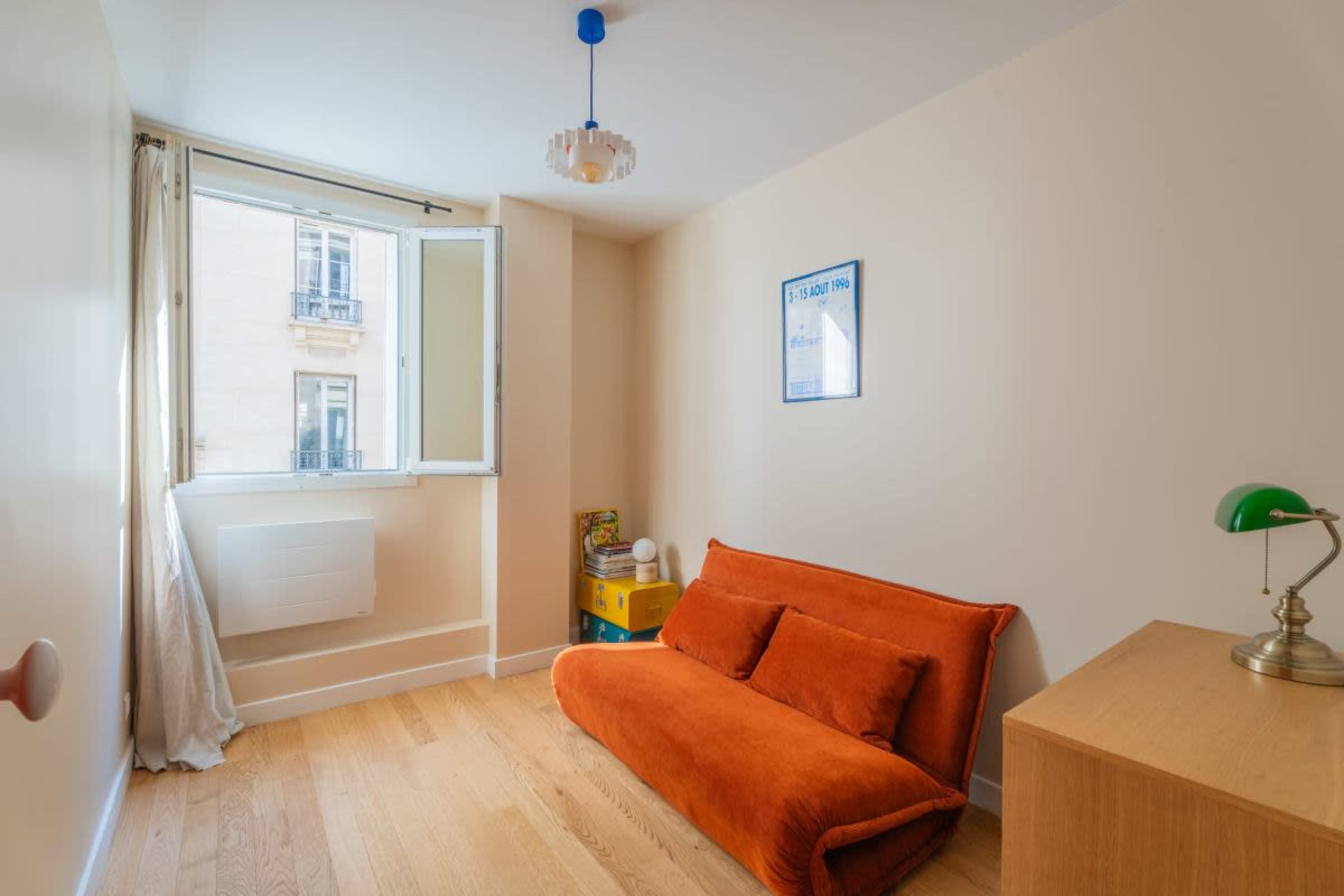 A small, well-lit room features an orange futon, a wooden nightstand, and a window that opens to a view of a building across the street.