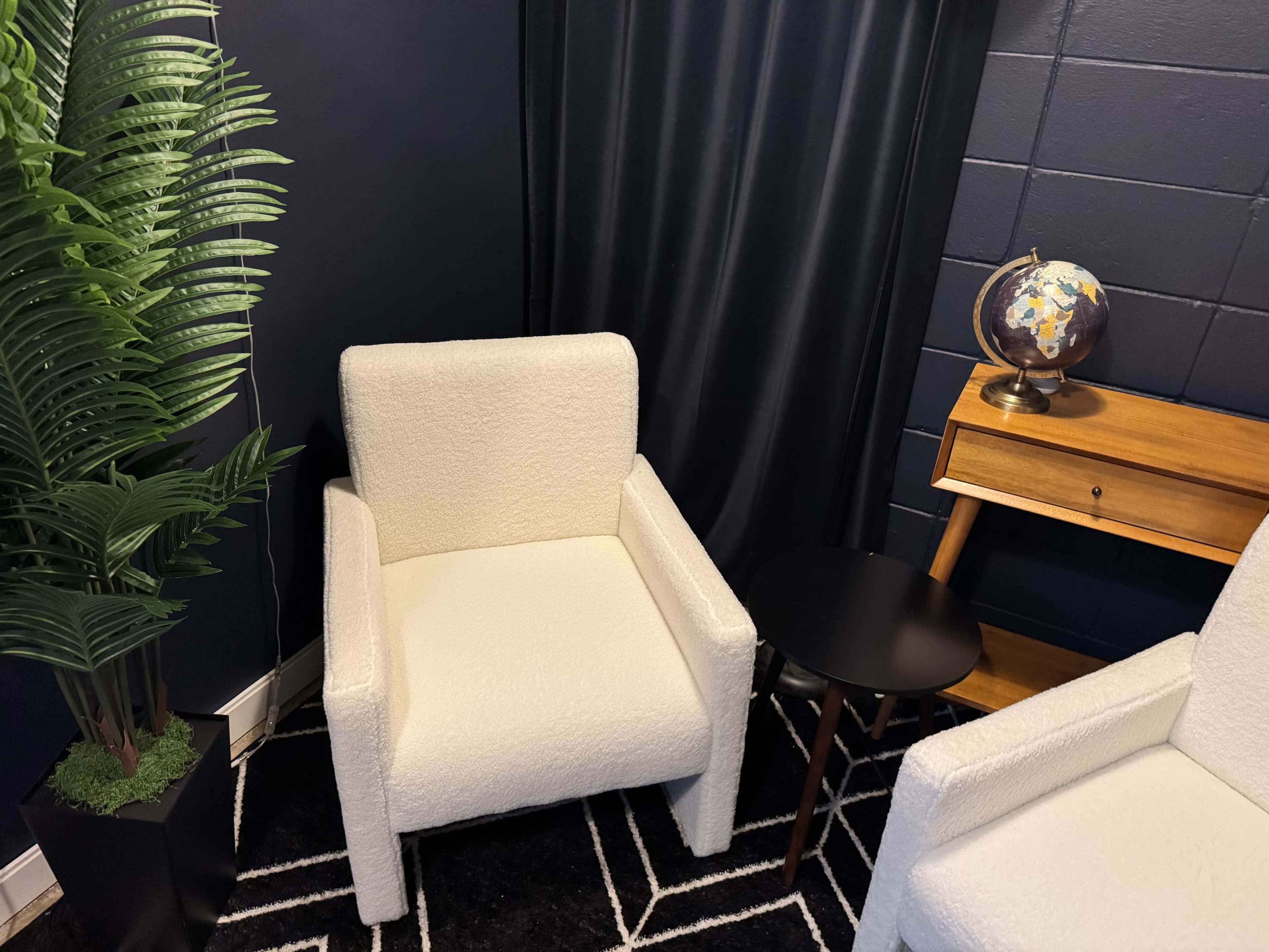 The image shows a corner of a room featuring two white chairs, a small black side table, a wooden desk with a globe, and a tall green plant against a dark wall.