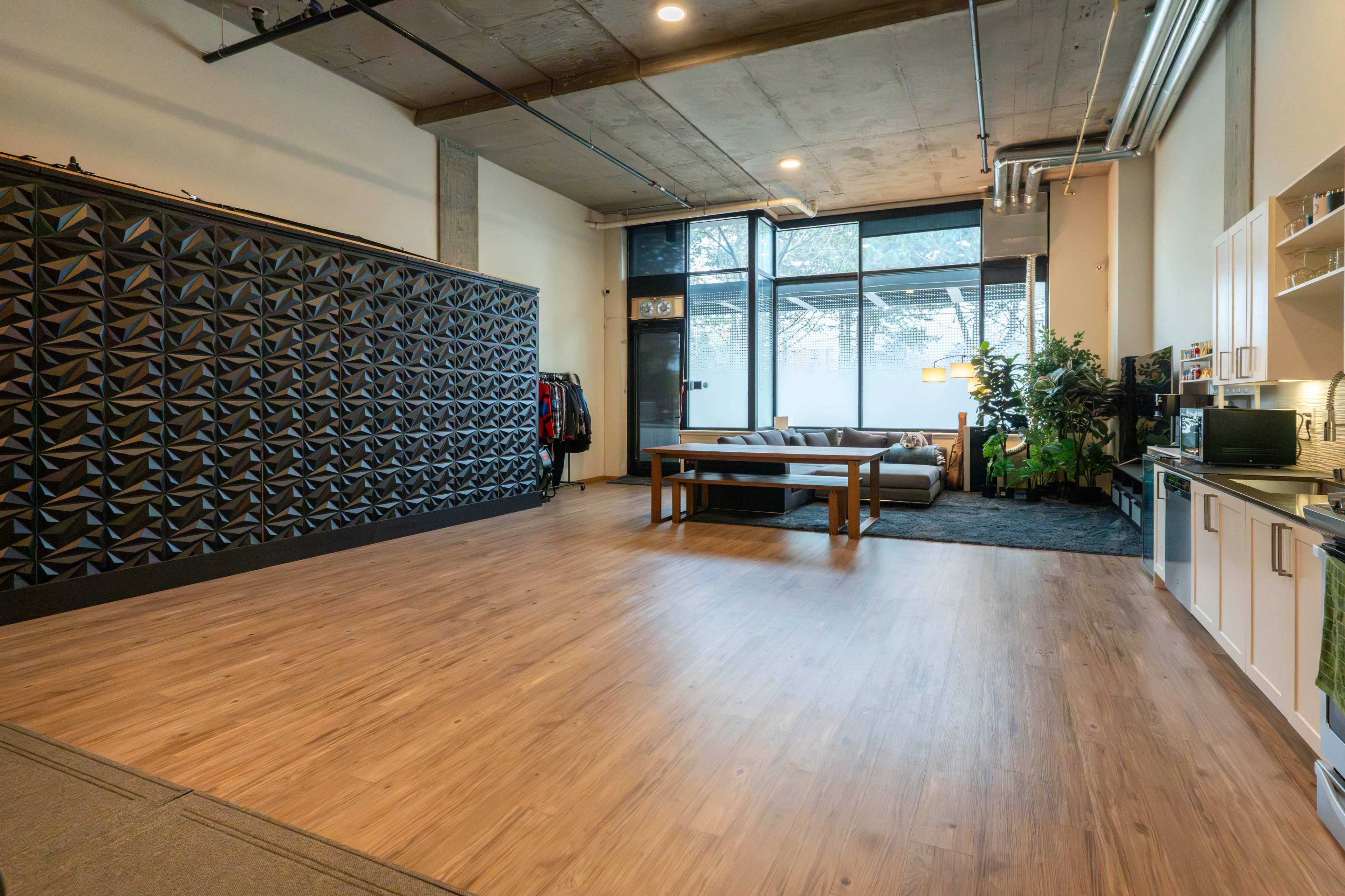 The image shows a spacious, modern interior with wood flooring, large windows, and a decorative wall with a geometric pattern.
