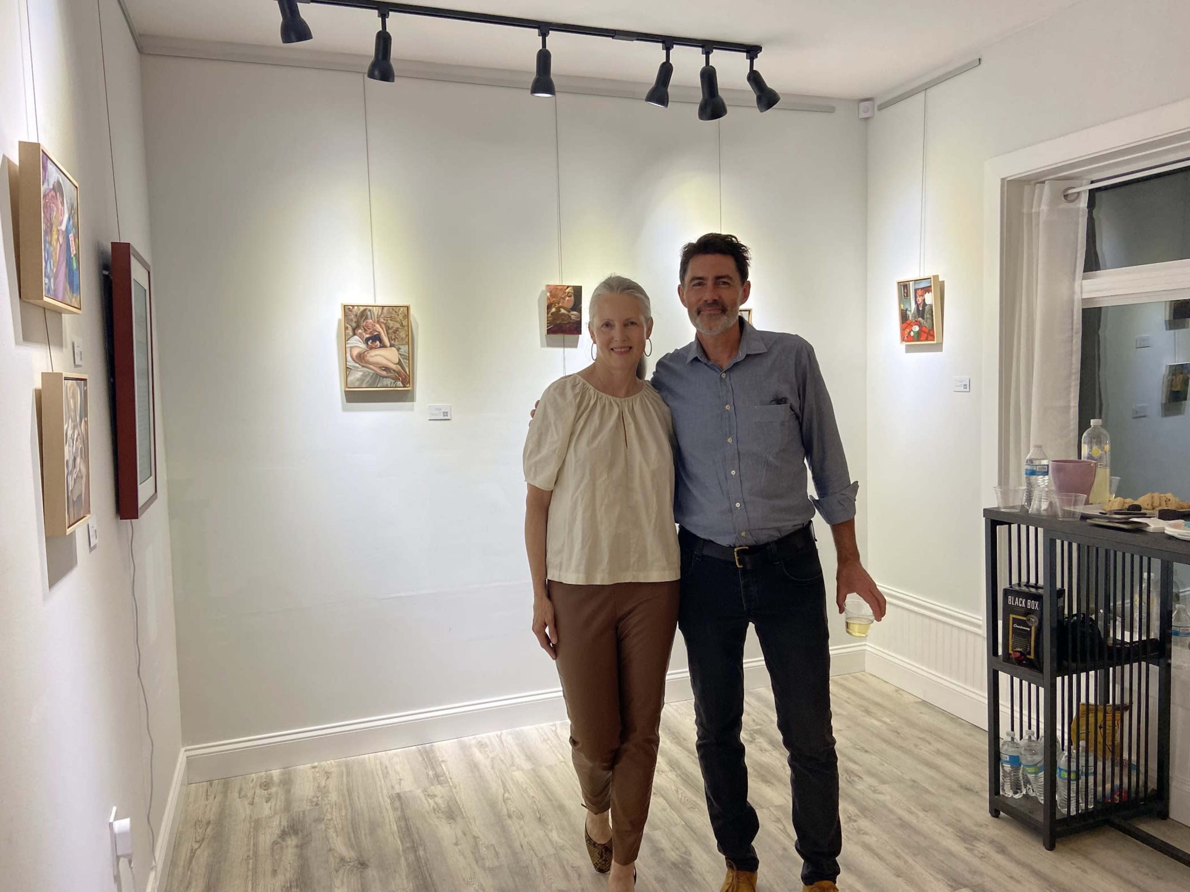 A man and a woman stand together in an art gallery, surrounded by framed artworks on the walls.