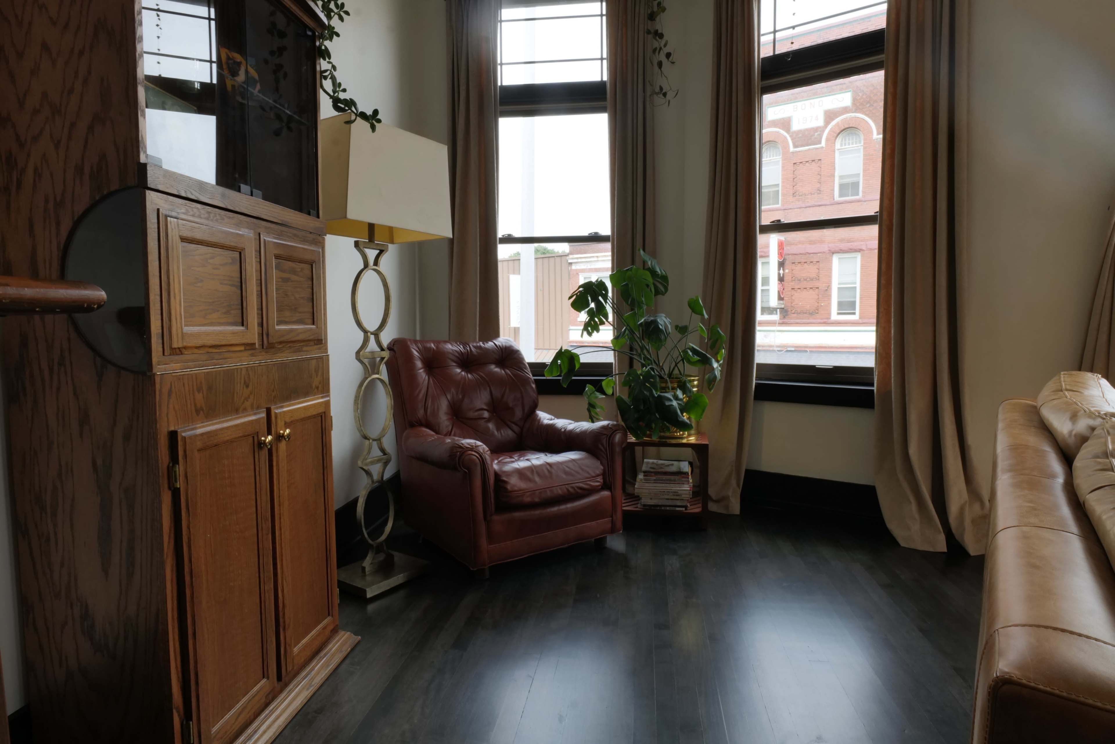 The image shows a corner of a room featuring a brown leather armchair, a potted plant, a tall lamp, and large windows with curtains.