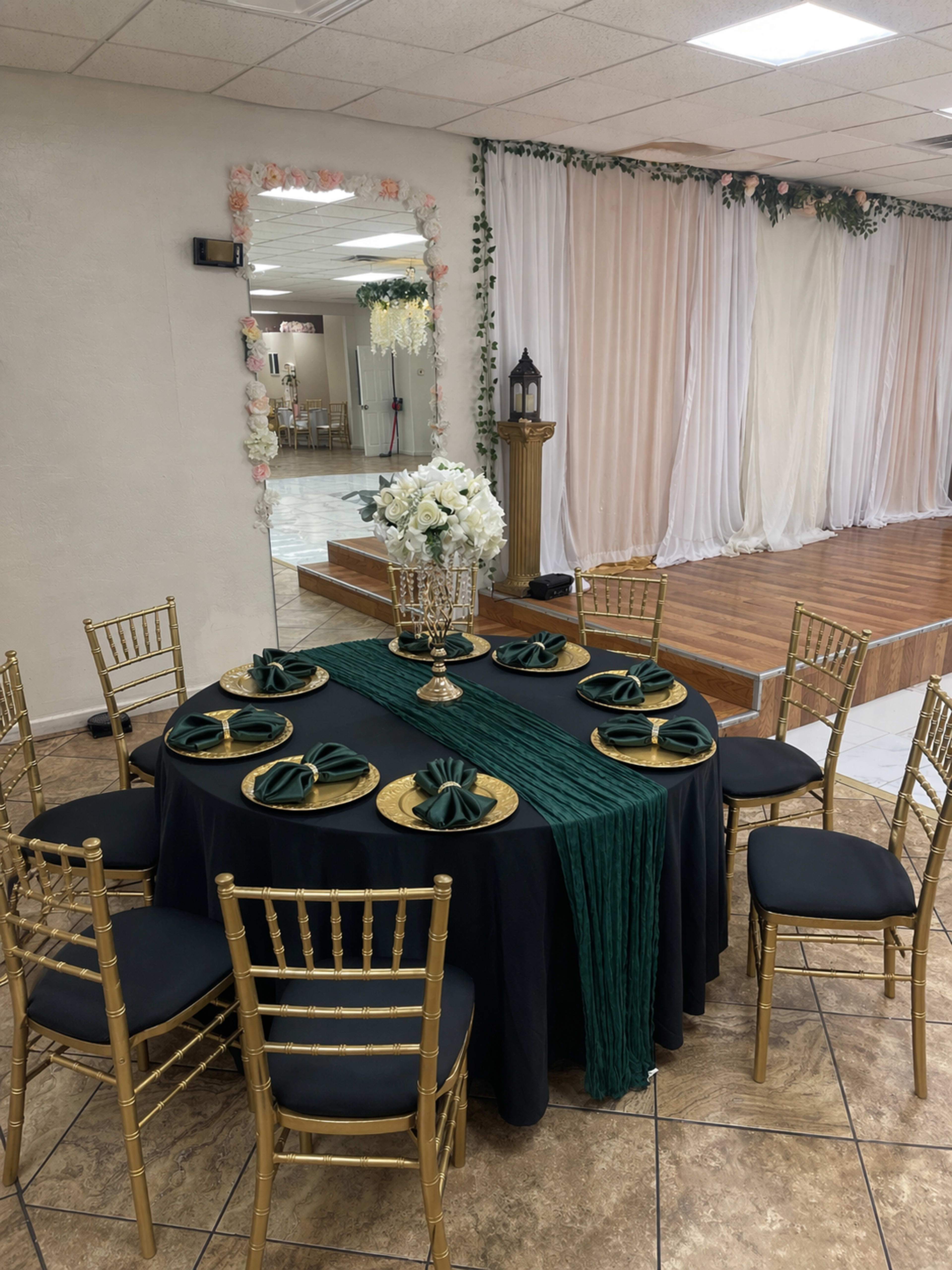 A round dining table set with green napkins and a floral center arrangement is surrounded by gold chairs in a banquet hall.