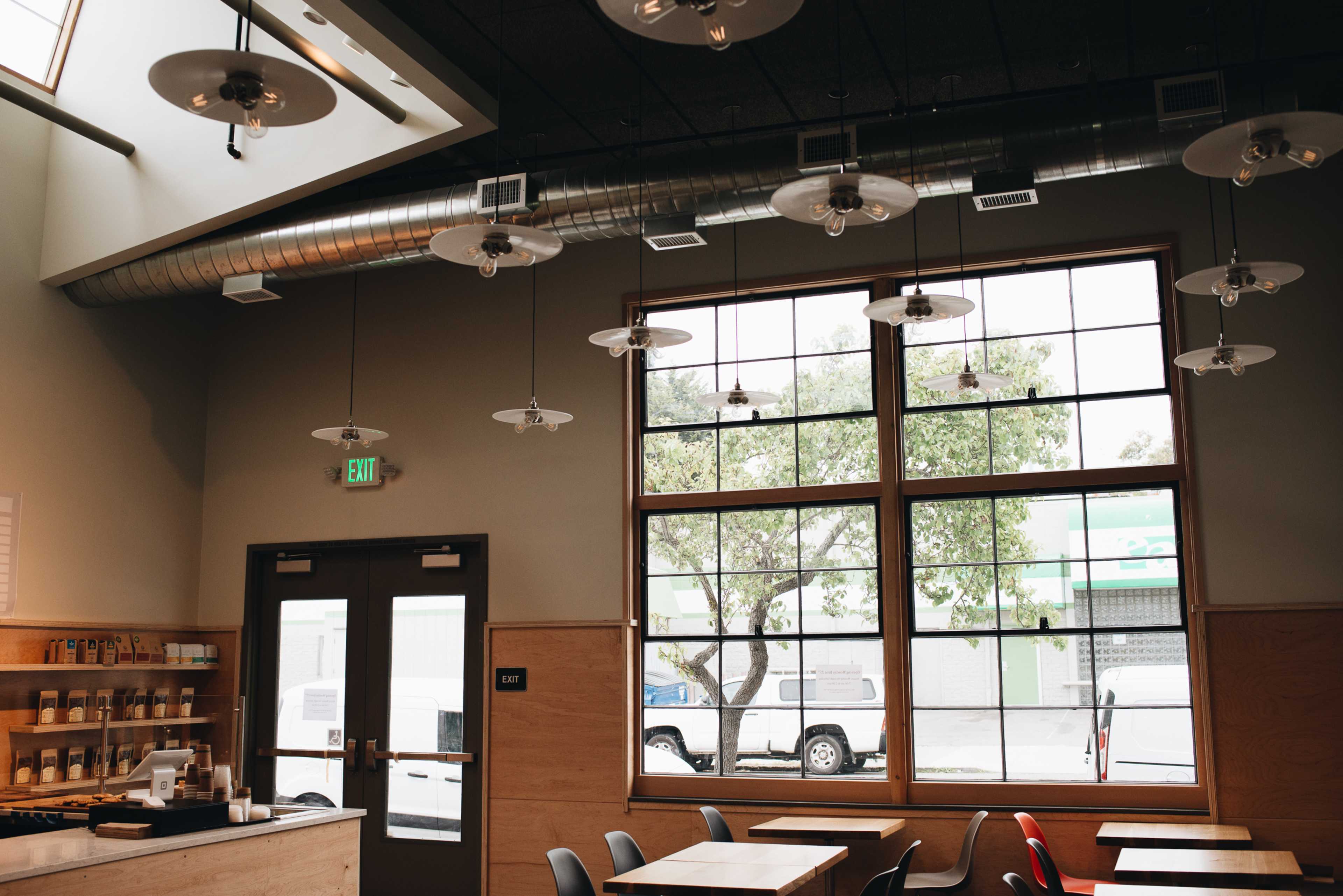 The interior of a café features large windows, wooden tables, and industrial-style light fixtures hanging from the ceiling.