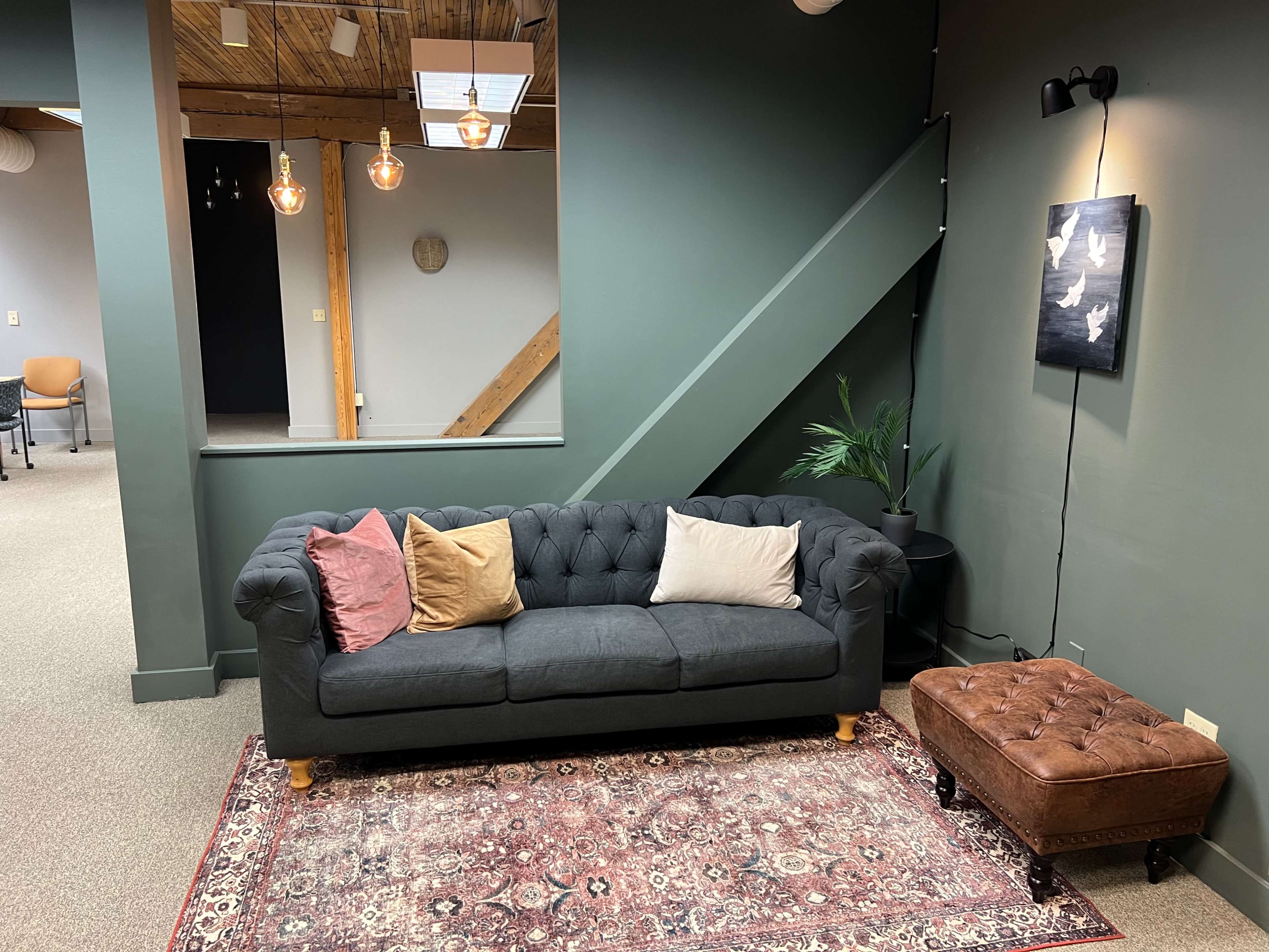 A cozy seating area with a tufted gray sofa, decorative pillows, a tufted ottoman, and a potted plant, situated beneath a staircase in a well-lit room.