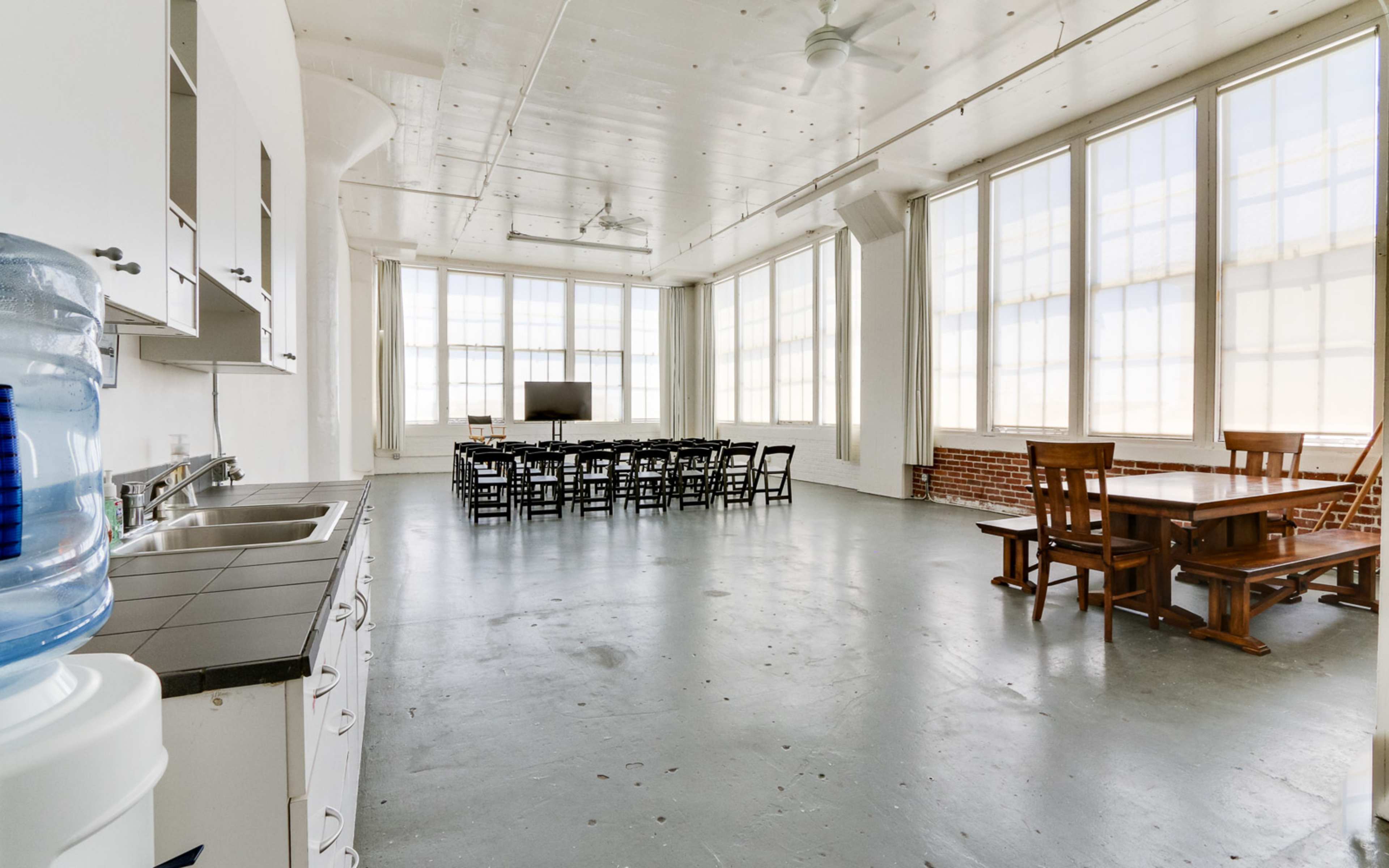 A spacious, well-lit room with large windows, featuring a kitchen area, a long dining table, and a separate seating area.
