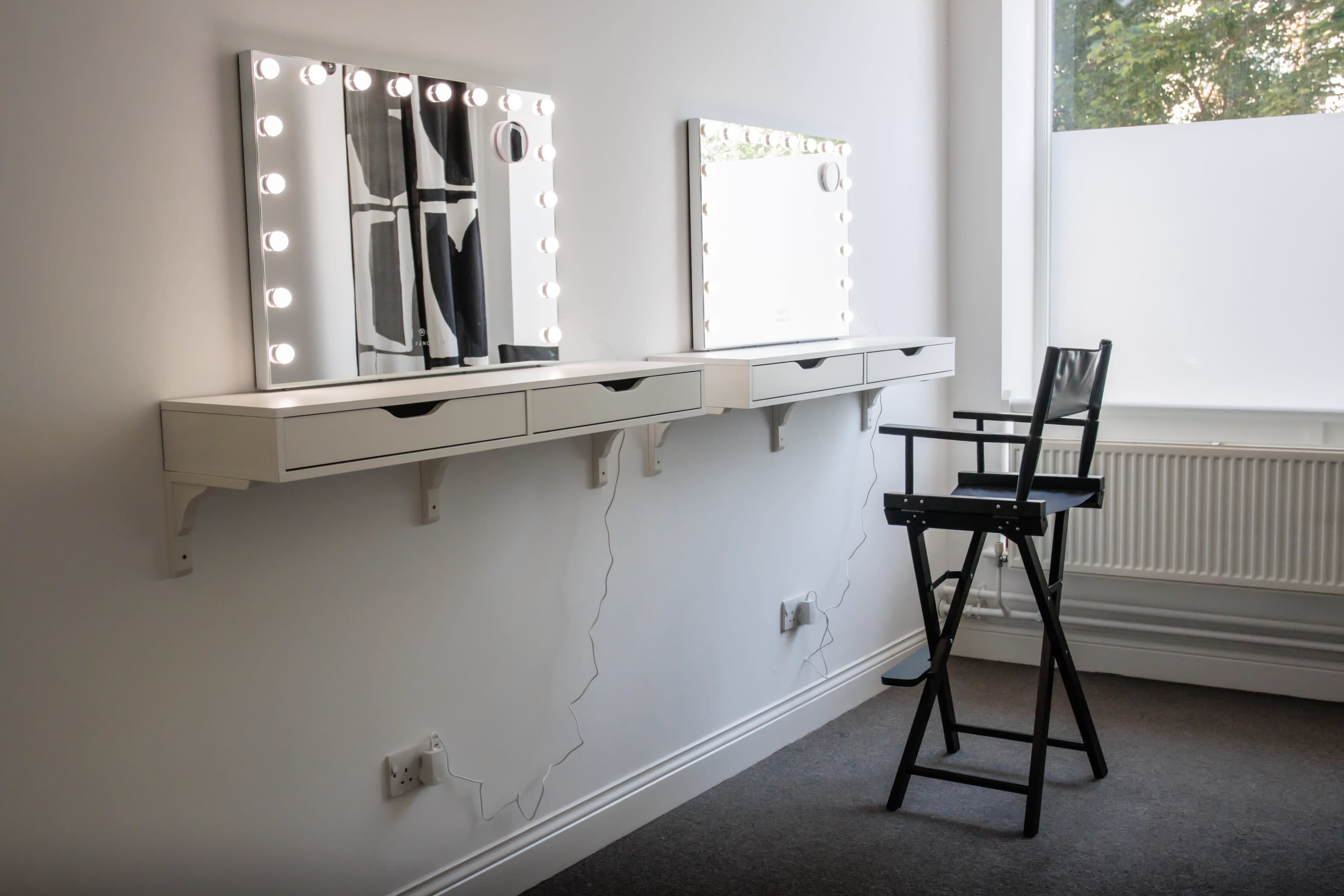 A well-lit makeup station with two mirrors and a black director's chair against a white wall.