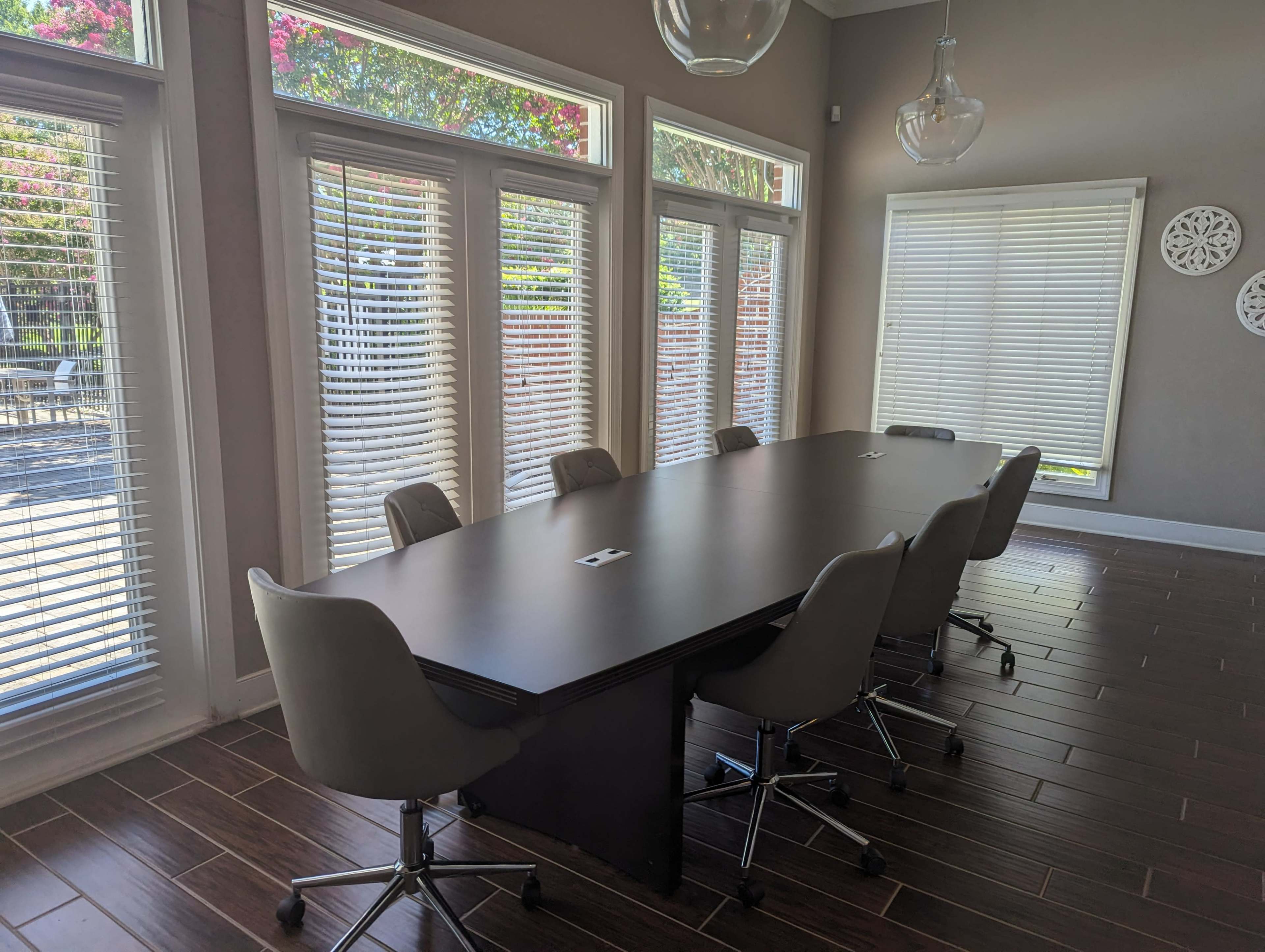 A long conference table with rolling chairs is positioned in a brightly lit room featuring large windows and blinds.