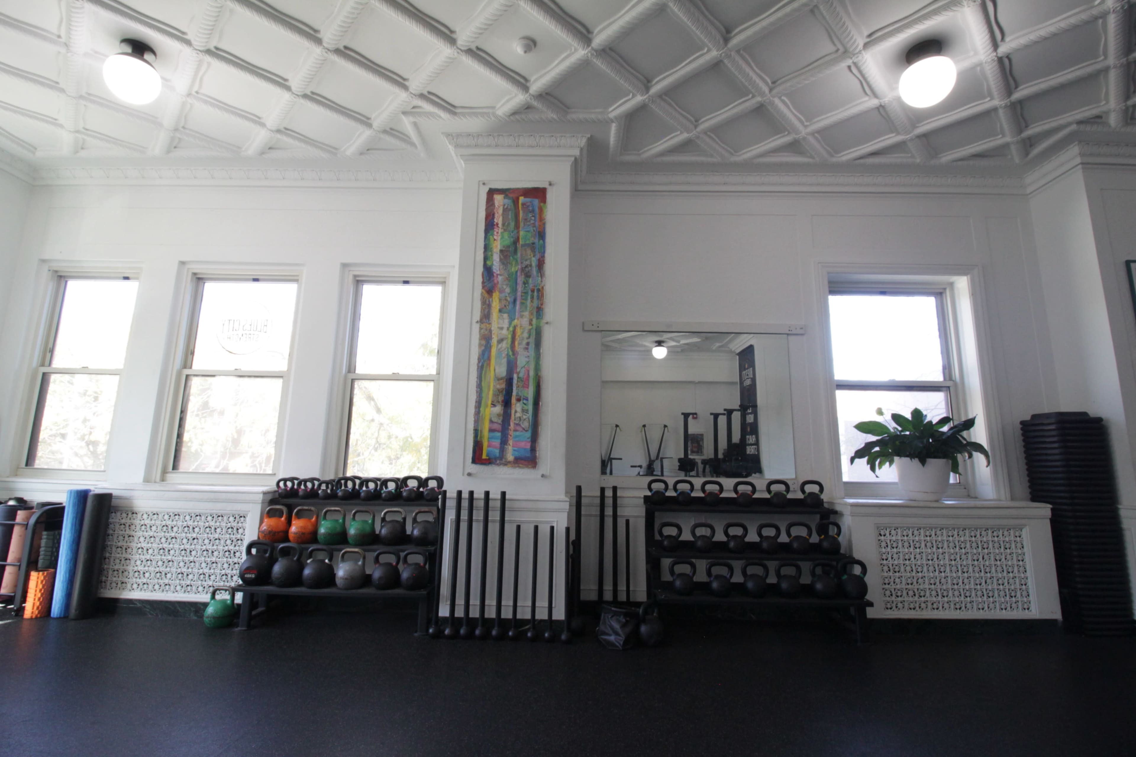 The image shows a fitness studio with kettlebells and weights neatly arranged on shelves, large windows letting in natural light, and a decorative wall piece.