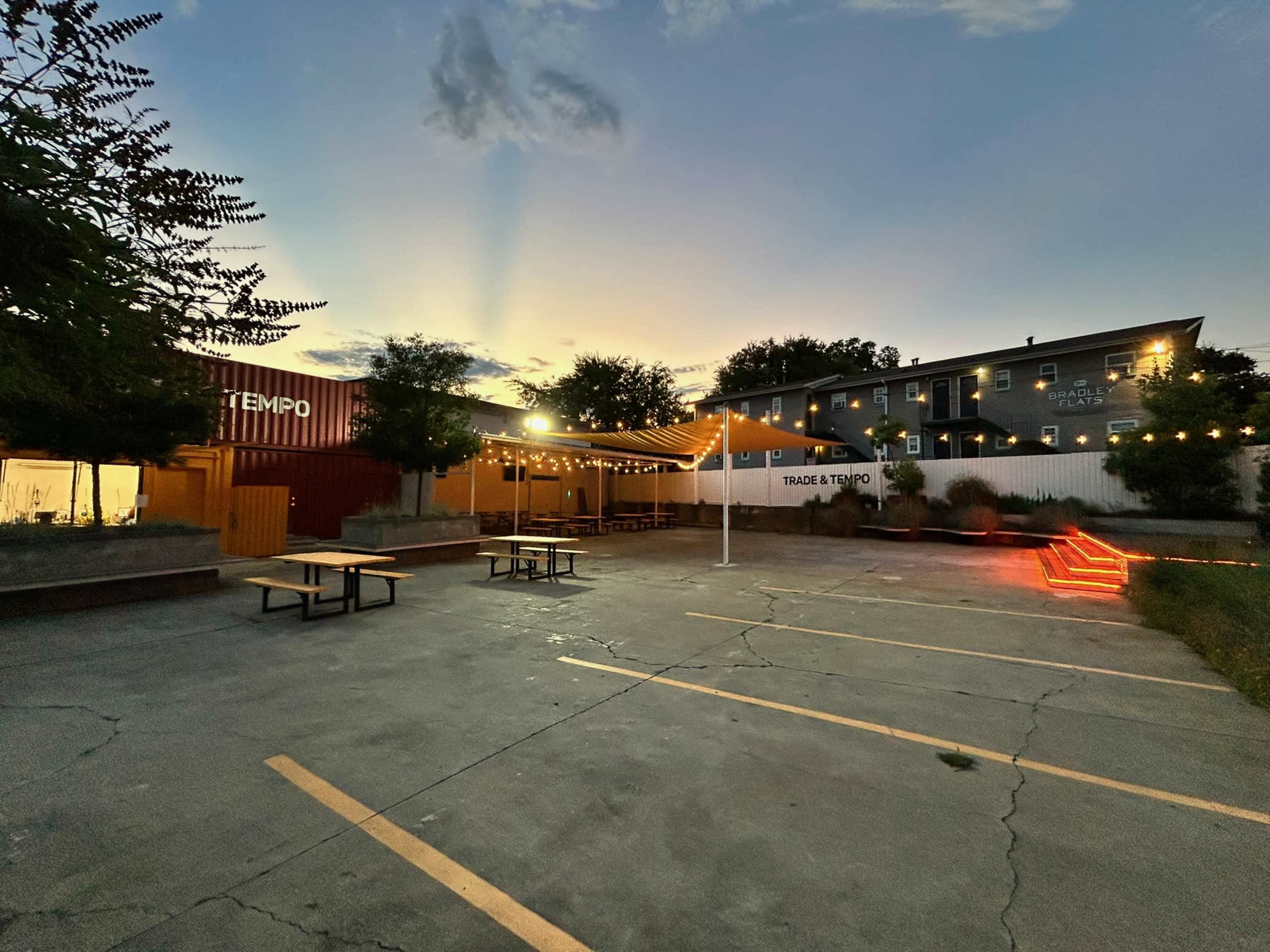 The image shows an outdoor area with seating, string lights, and a grassy border, set against a backdrop of a sunset and nearby buildings.