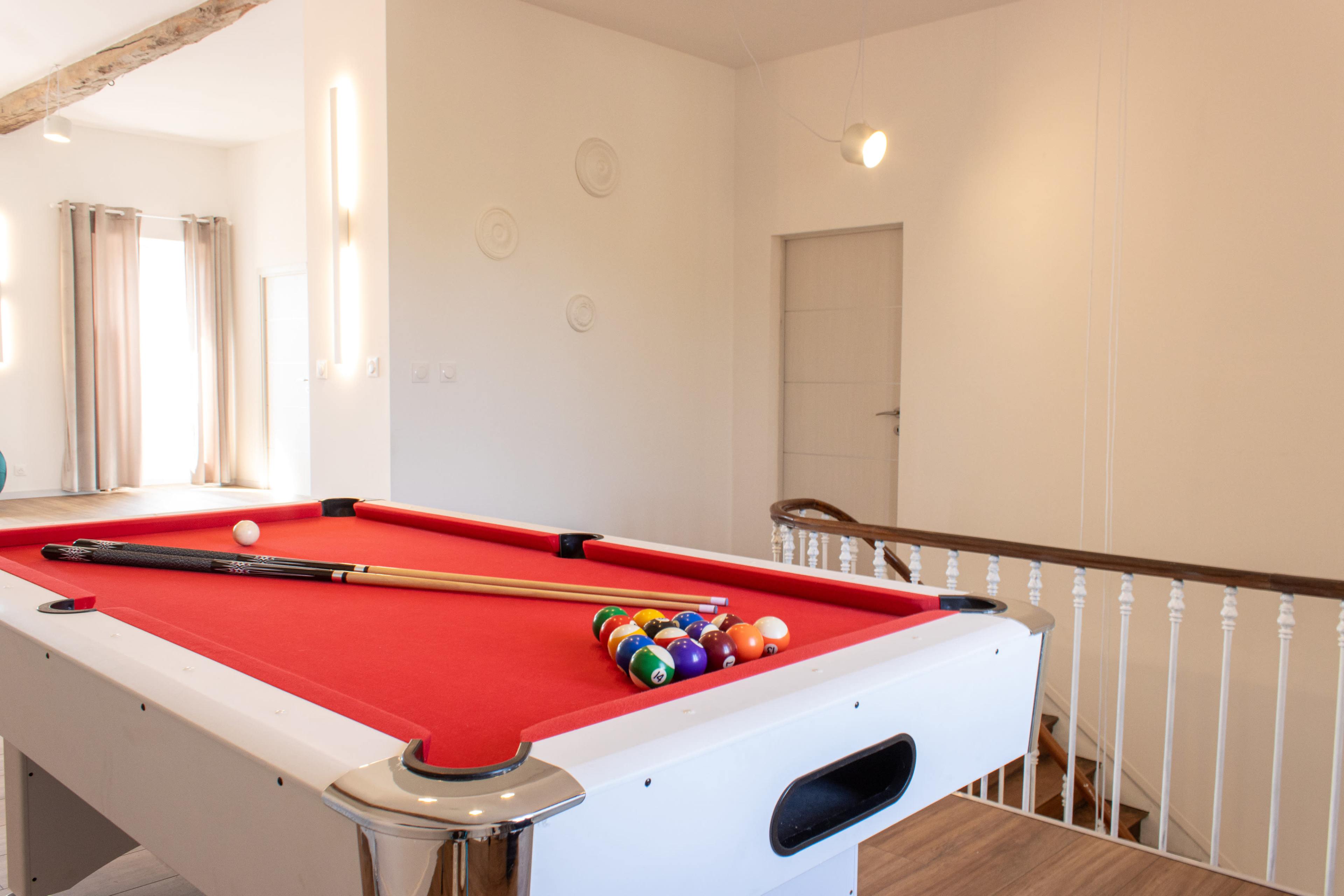A pool table with a red felt surface and a set of balls is positioned near a staircase in a brightly lit room.