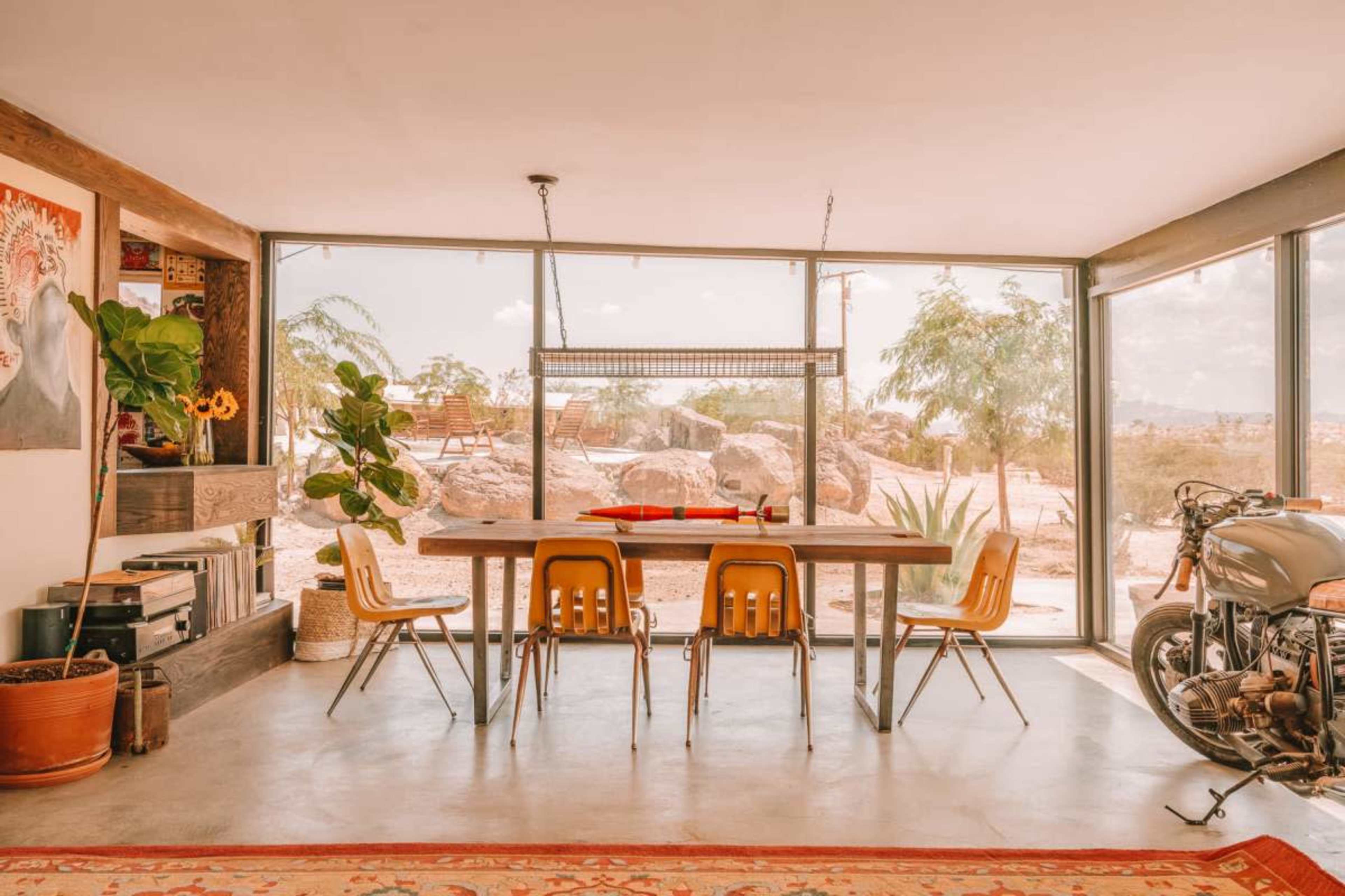 A dining area features a long wooden table surrounded by yellow chairs, with large windows showcasing a desert landscape.