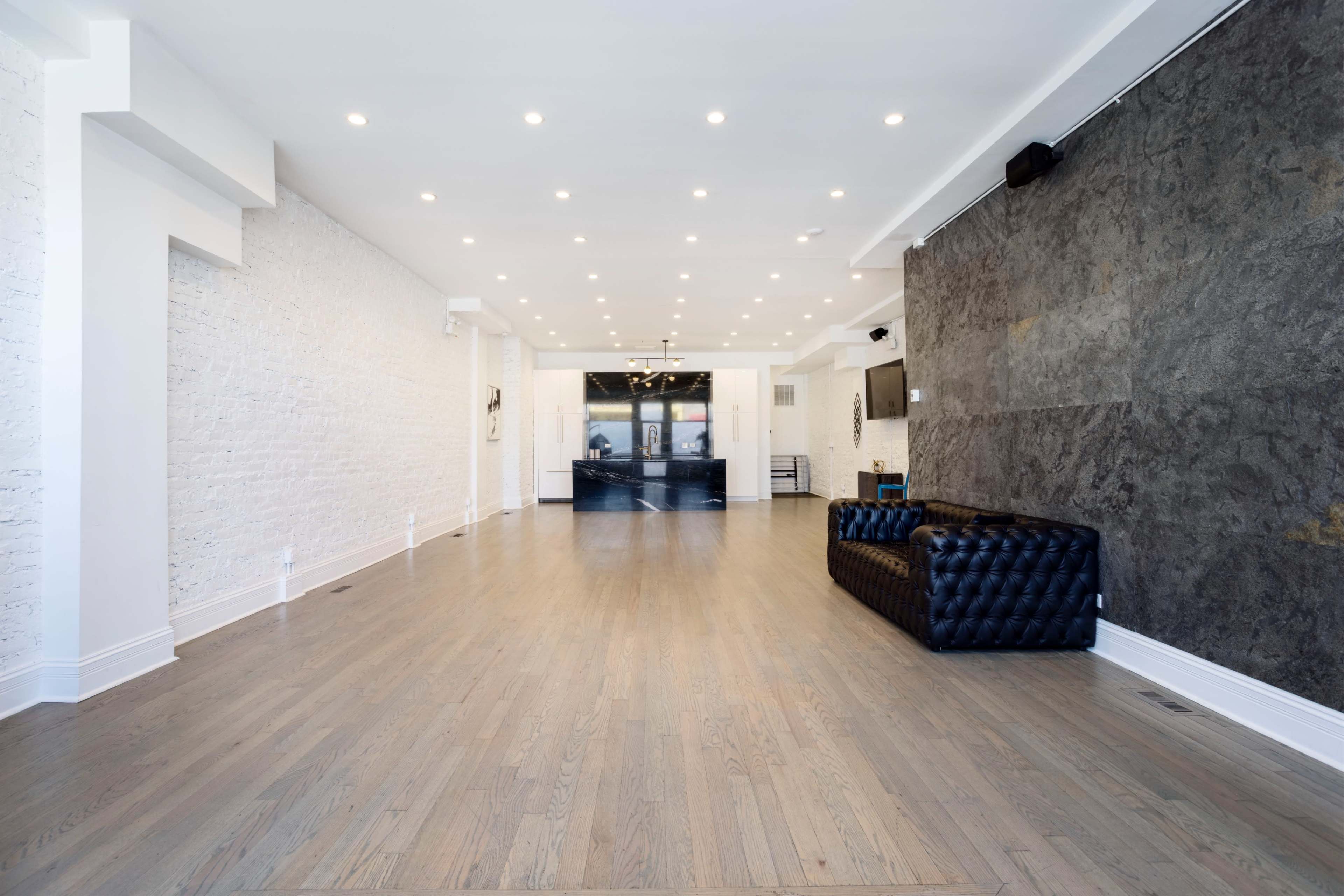 A spacious interior with light wood flooring, white brick walls, and a dark textured accent wall, featuring a stylish black couch and a counter in the background.