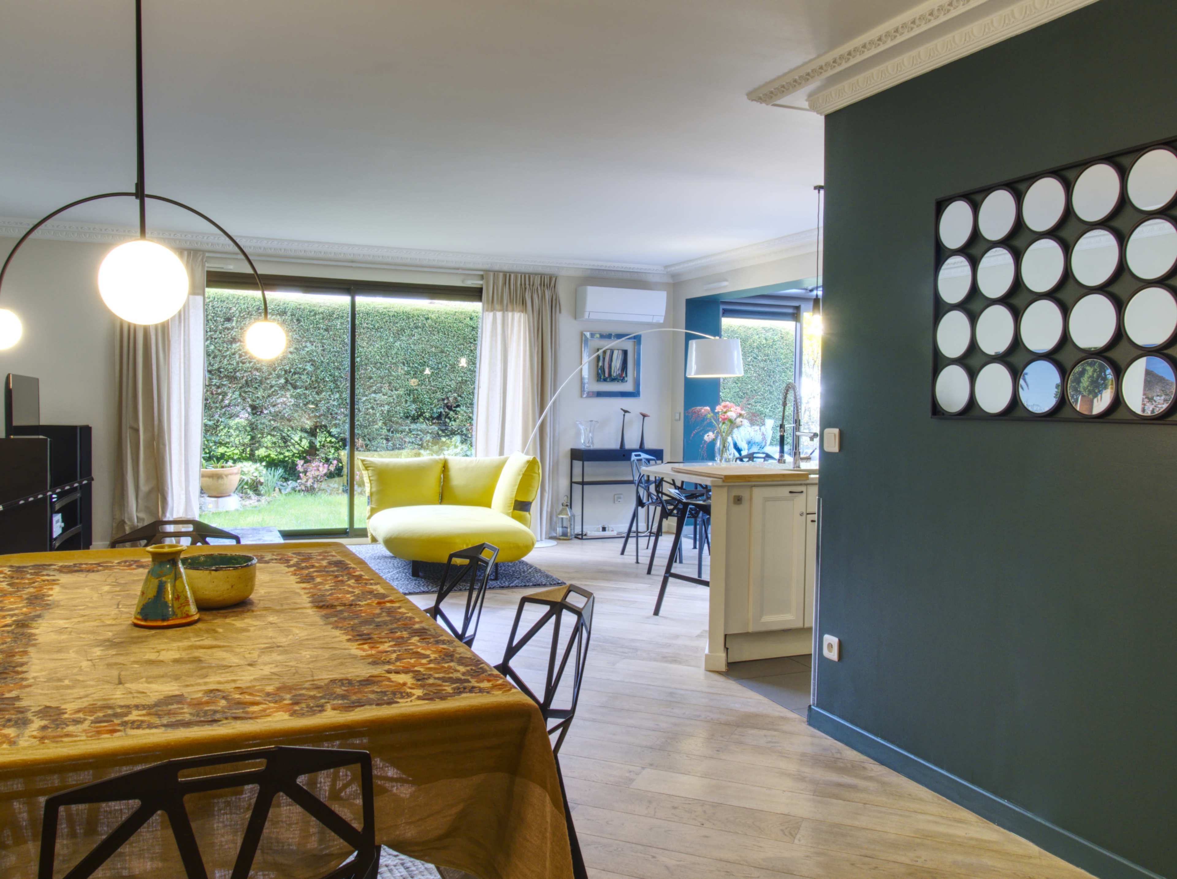 The image shows a modern living space featuring a dining area with a patterned tablecloth, a bright green sofa, and large windows opening to a garden.